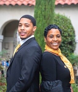 Two people stand back-to-back outside. One in a suit, the other in a black top with gold scarf.