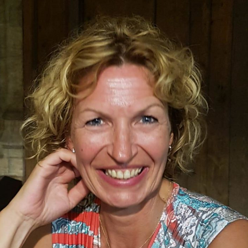 Een vrouw met krullend blond haar lacht naar de camera, terwijl ze een topje met een patroon draagt.