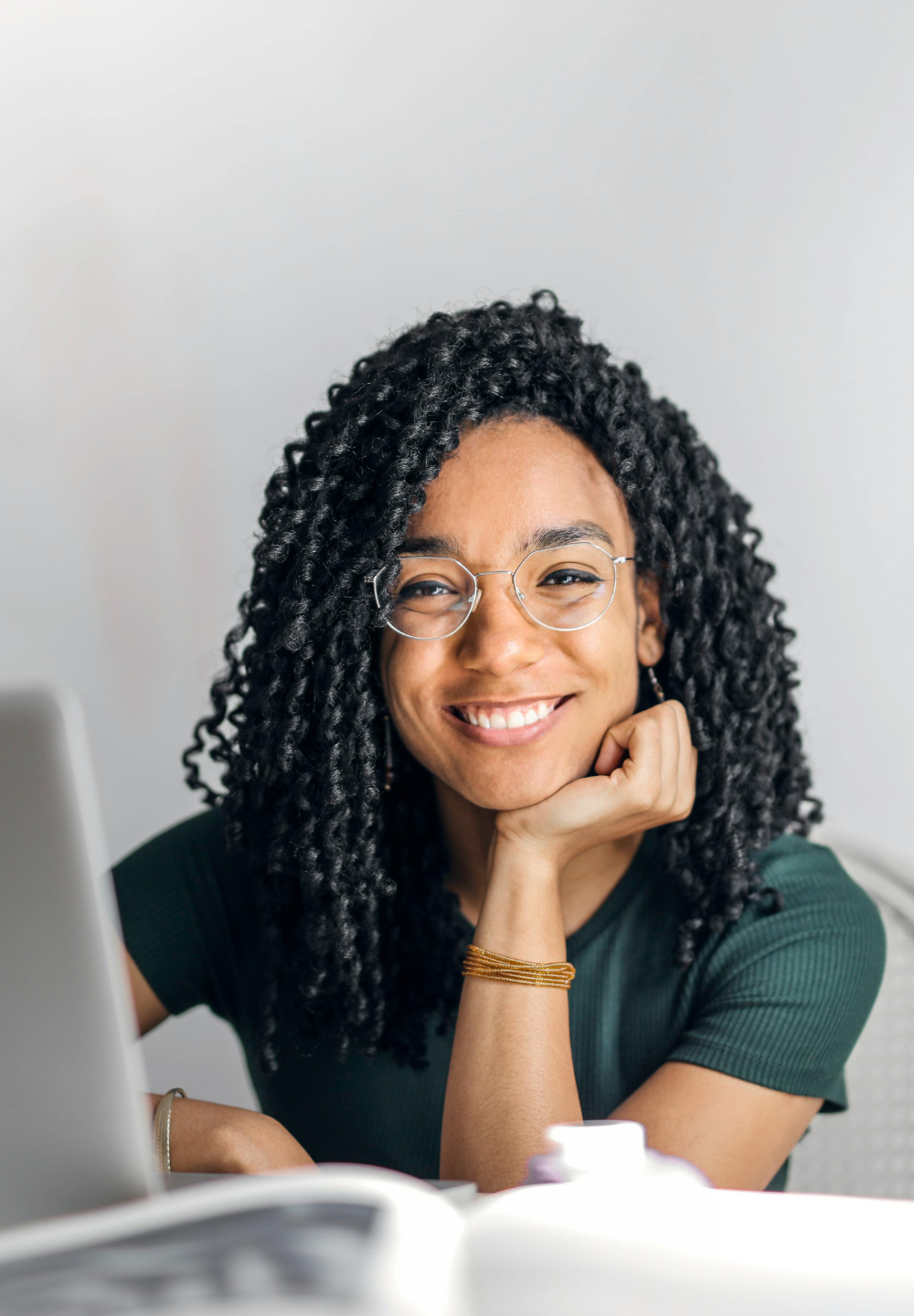Een vrouw met krullend donker haar en een bril lacht naar de camera, terwijl ze haar kin op haar hand laat rusten aan een bureau met een laptop en een open boek.