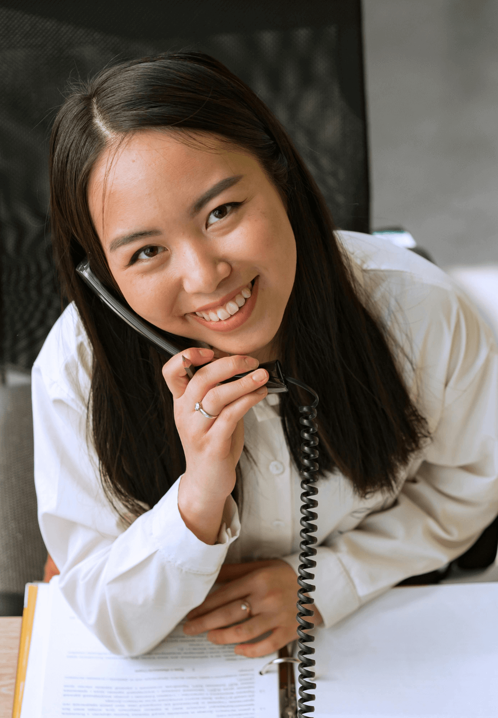 Vrouw leunend tegen een bureau, glimlachend terwijl ze een telefoon vasthoudt. Witte blouse, lang donker haar, kantooromgeving.
