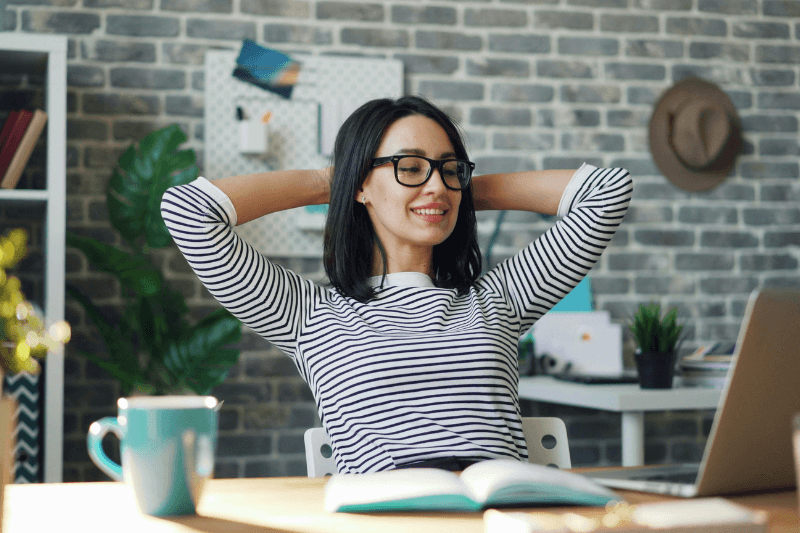 Vrouw met bril glimlacht, ontspannen zittend aan een bureau met laptop en boek; bakstenen muur op de achtergrond.