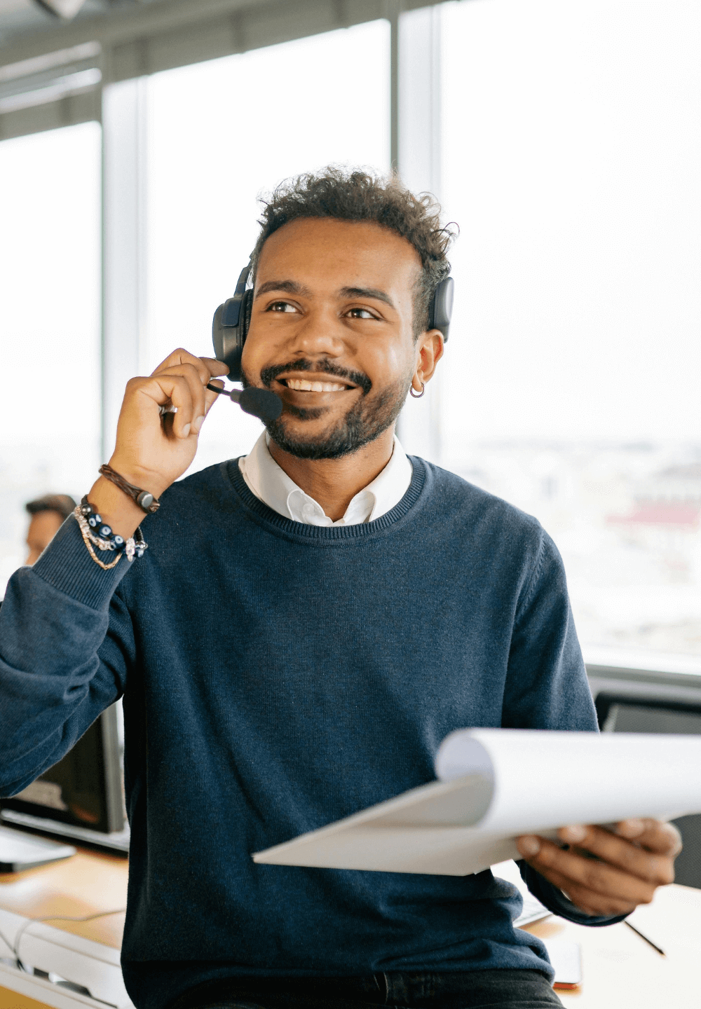 Een man met een headset glimlacht, houdt papieren vast en kijkt omhoog in een helder verlicht kantoor.