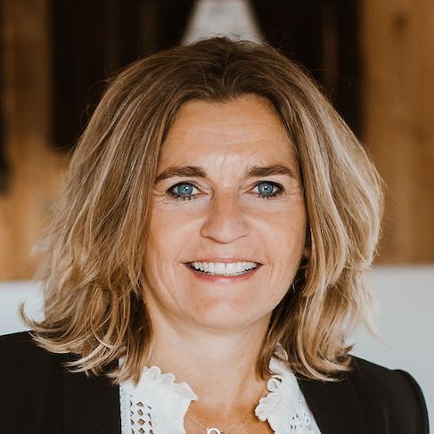 Vrouw met blond haar lacht, draagt ​​een zwarte blazer en een witte blouse, binnen.