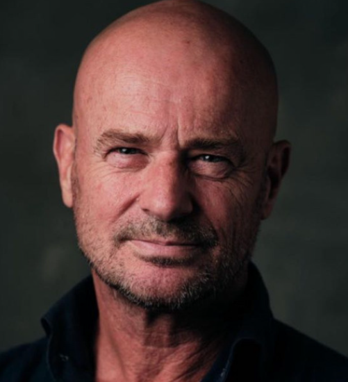 Een kale man met blauwe ogen en een lichte glimlach, gekleed in een blauw shirt, tegen een houten achtergrond.