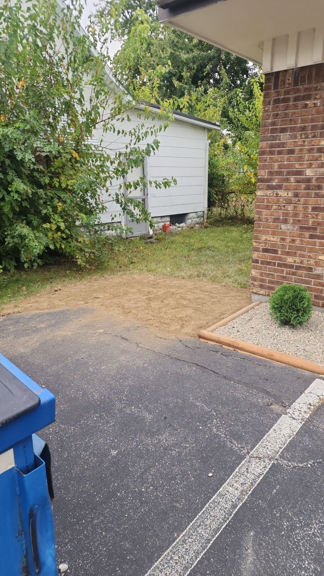 An asphalt driveway leads to a patch of straw, a white shed, and a brick house corner with a small shrub.