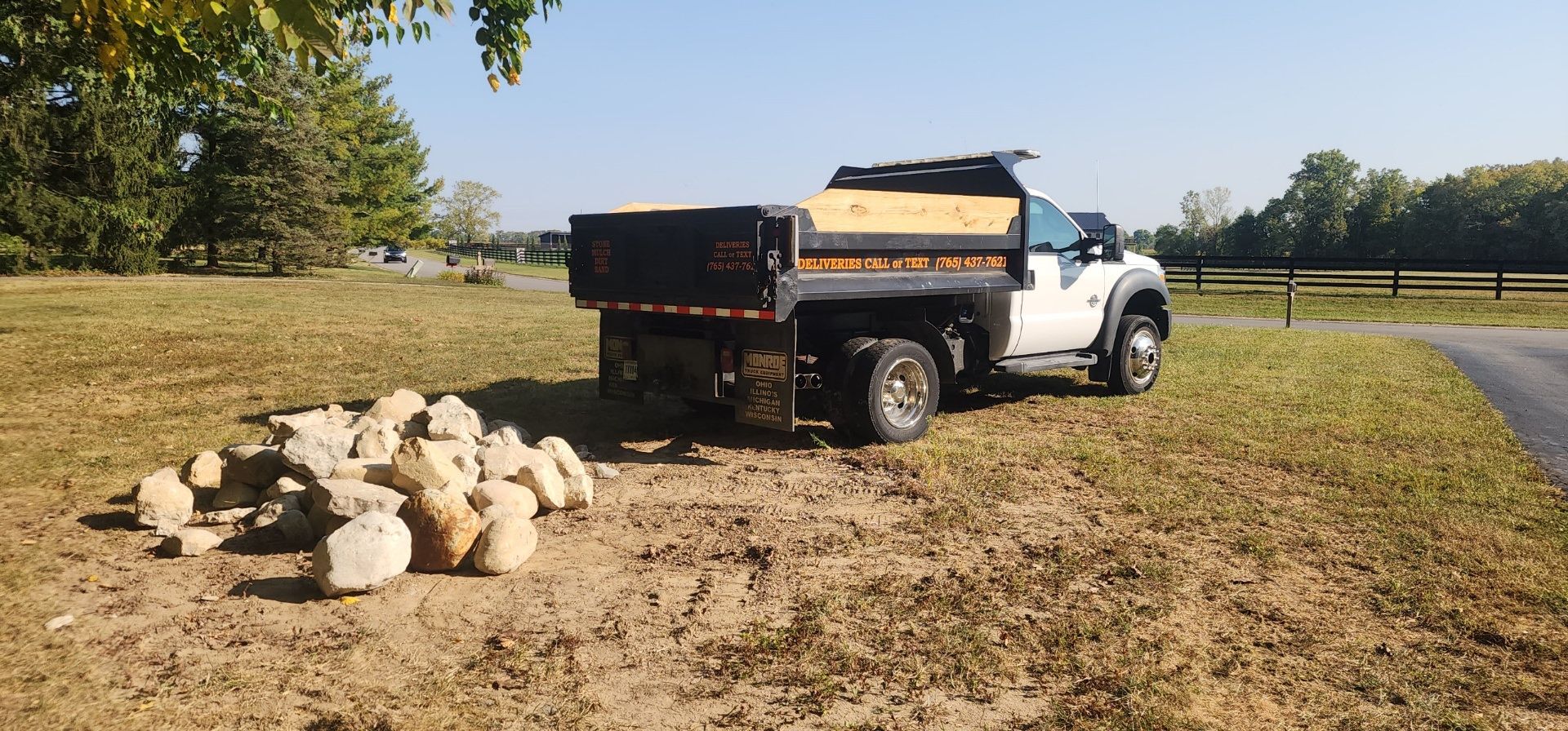 A white dump truck parked on a grassy field next to a small pile of light-colored boulders.
