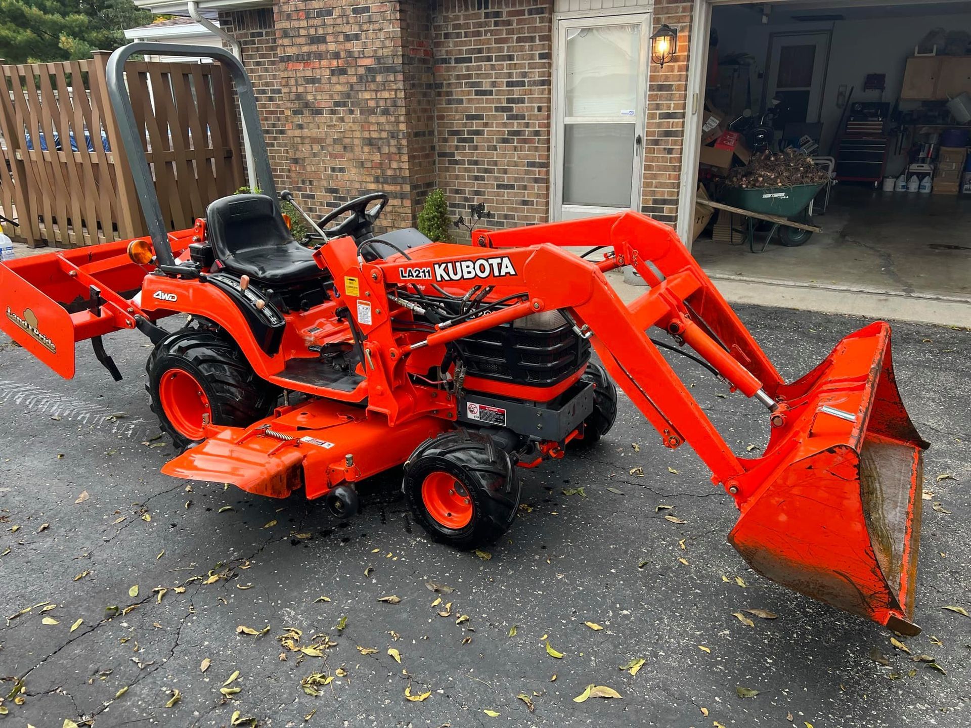 An orange tractor with a bucket attached to it is parked in front of a garage.