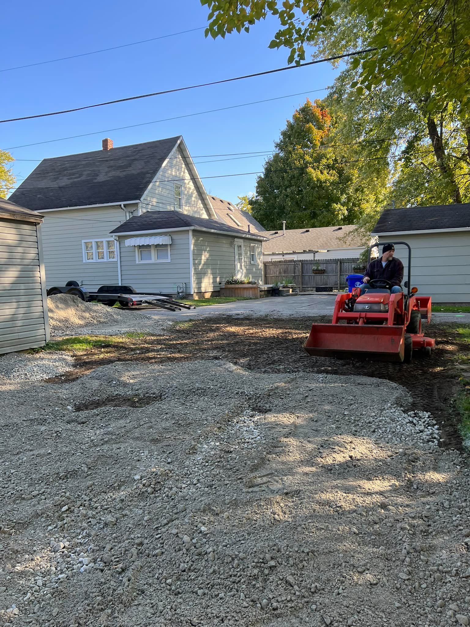 A man is driving a tractor in a gravel driveway in front of a house.