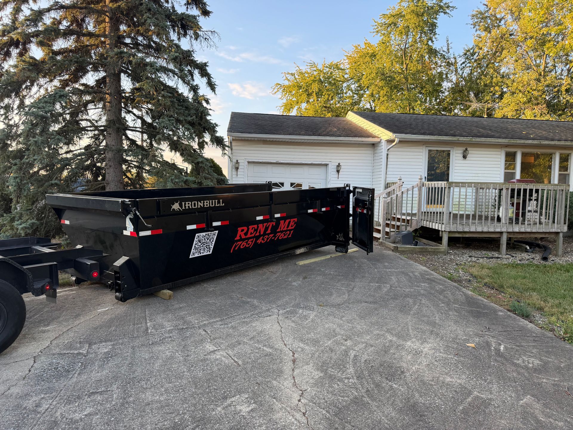 A large, black dumpster rental trailer sits on a concrete driveway in front of a white house with a deck.