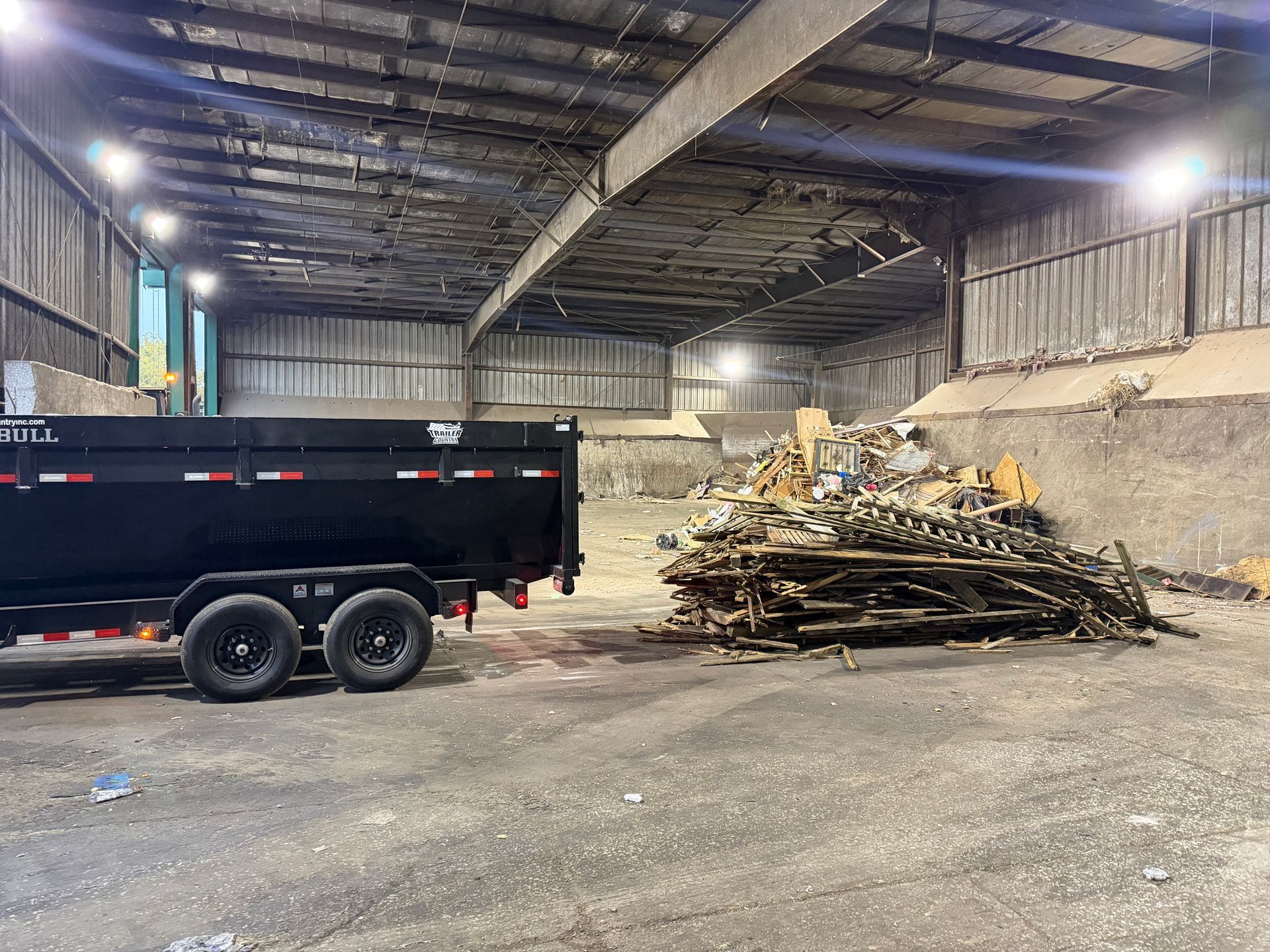 A black dump trailer parked inside a large, industrial warehouse next to a scattered pile of debris and scrap materials.