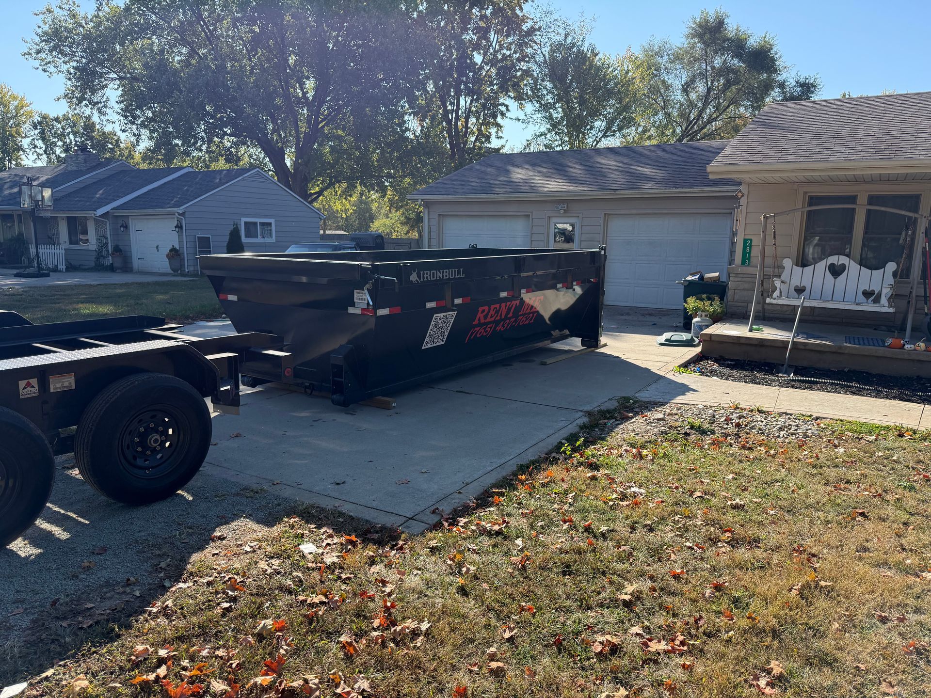 A black rental dumpster sits on a residential driveway next to a garage and house on a sunny day.
