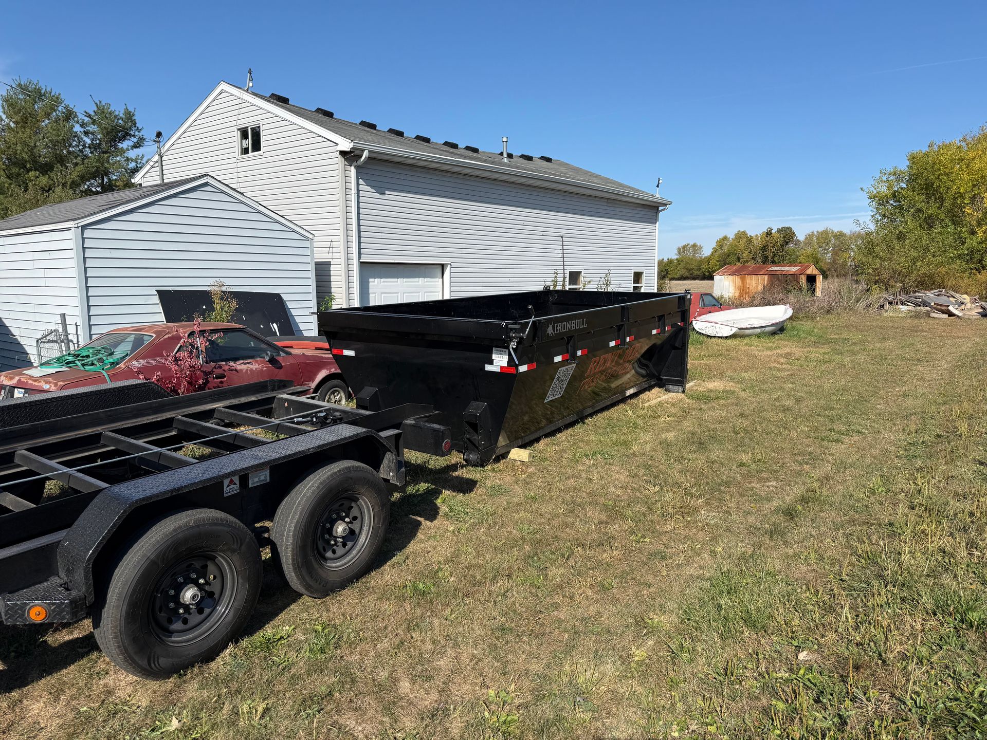 A black dump trailer sits attached to a flatbed trailer in a grassy lot next to a house and an old car.