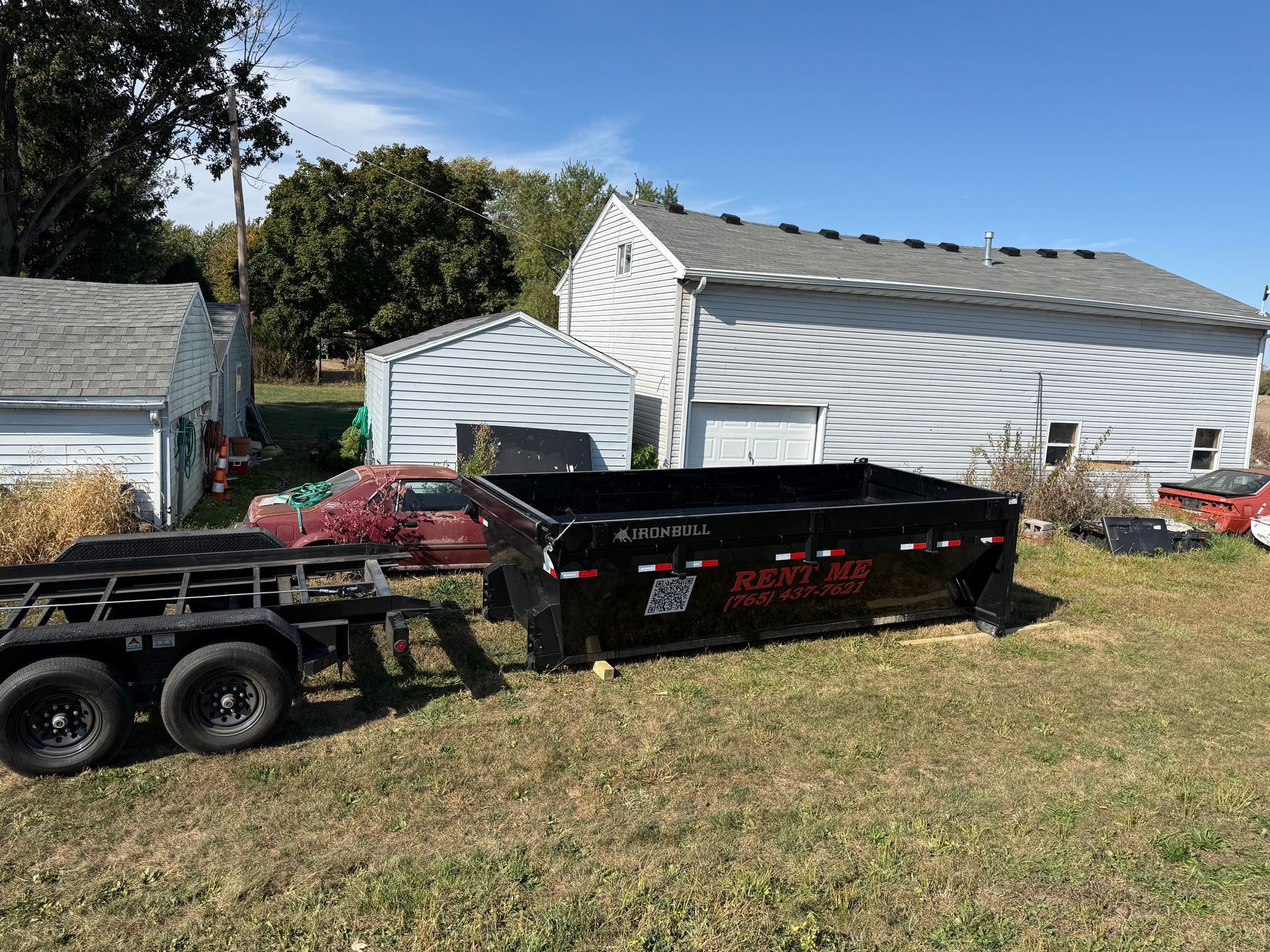 A large black dumpster, a flatbed trailer, and a red car parked in front of two white utility buildings on a grassy lot.
