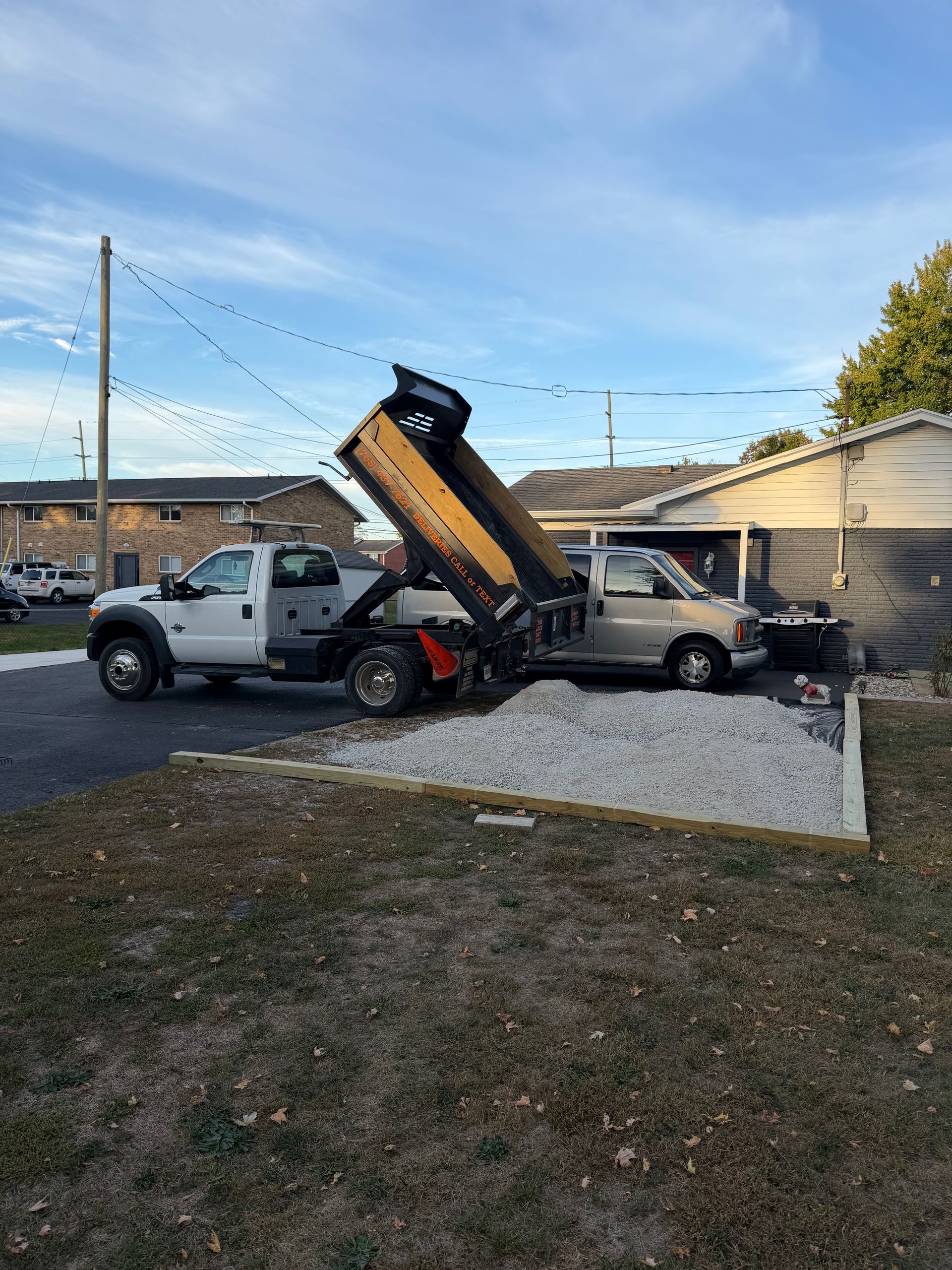 A white dump truck parked on a driveway with its bed raised, emptying a load of gravel onto the ground.