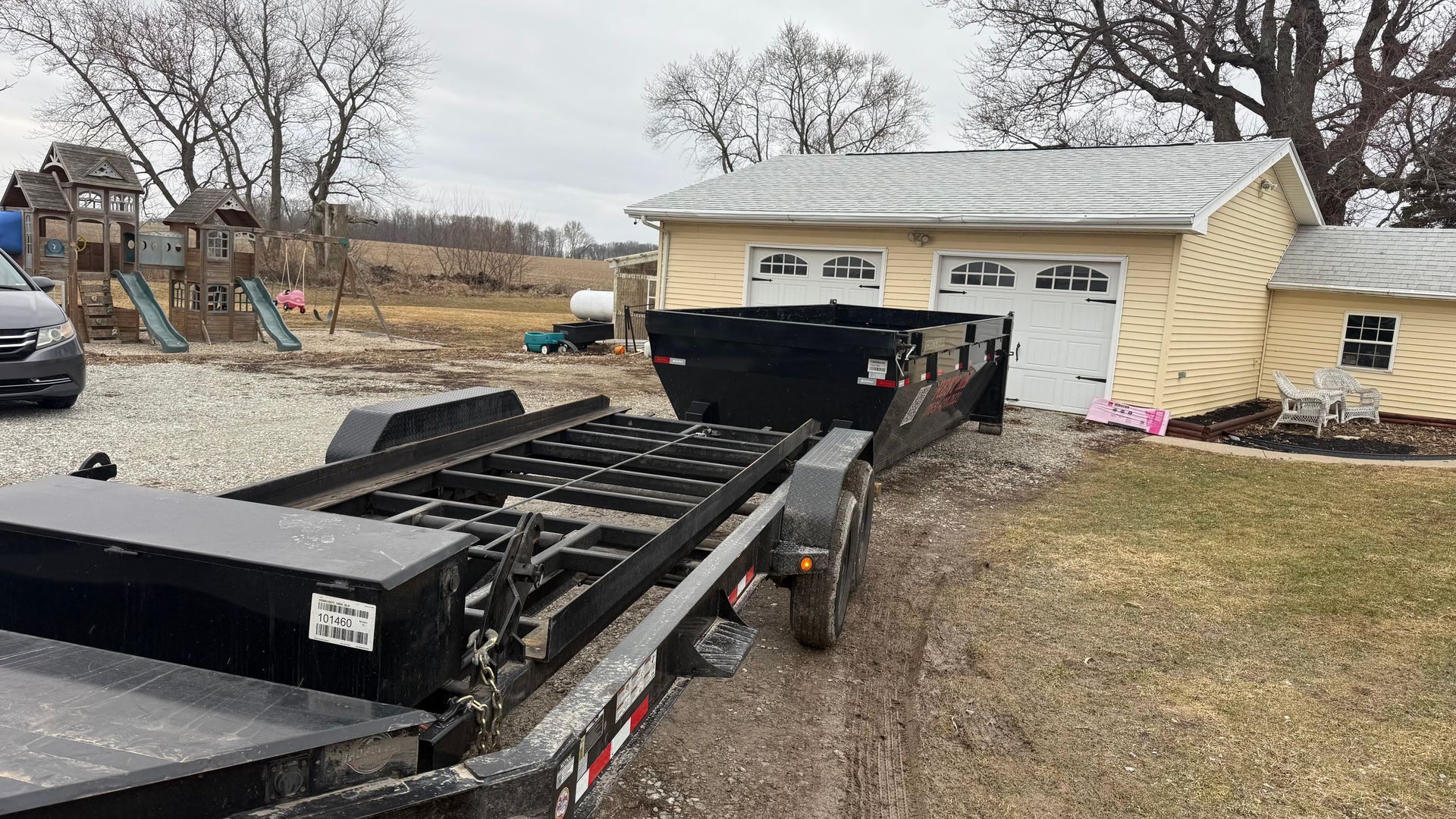 A black roll-off dumpster trailer parked on gravel near a garage, with a playground in the background.