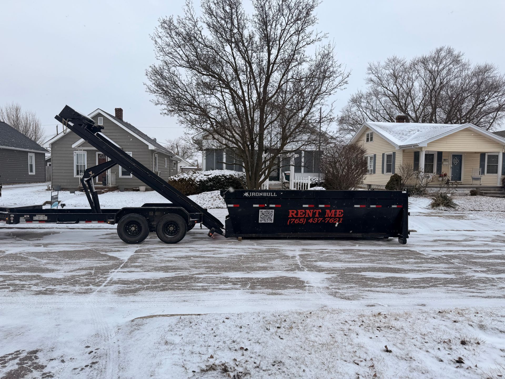 A black roll-off dumpster trailer sits on a snowy street in front of residential houses.