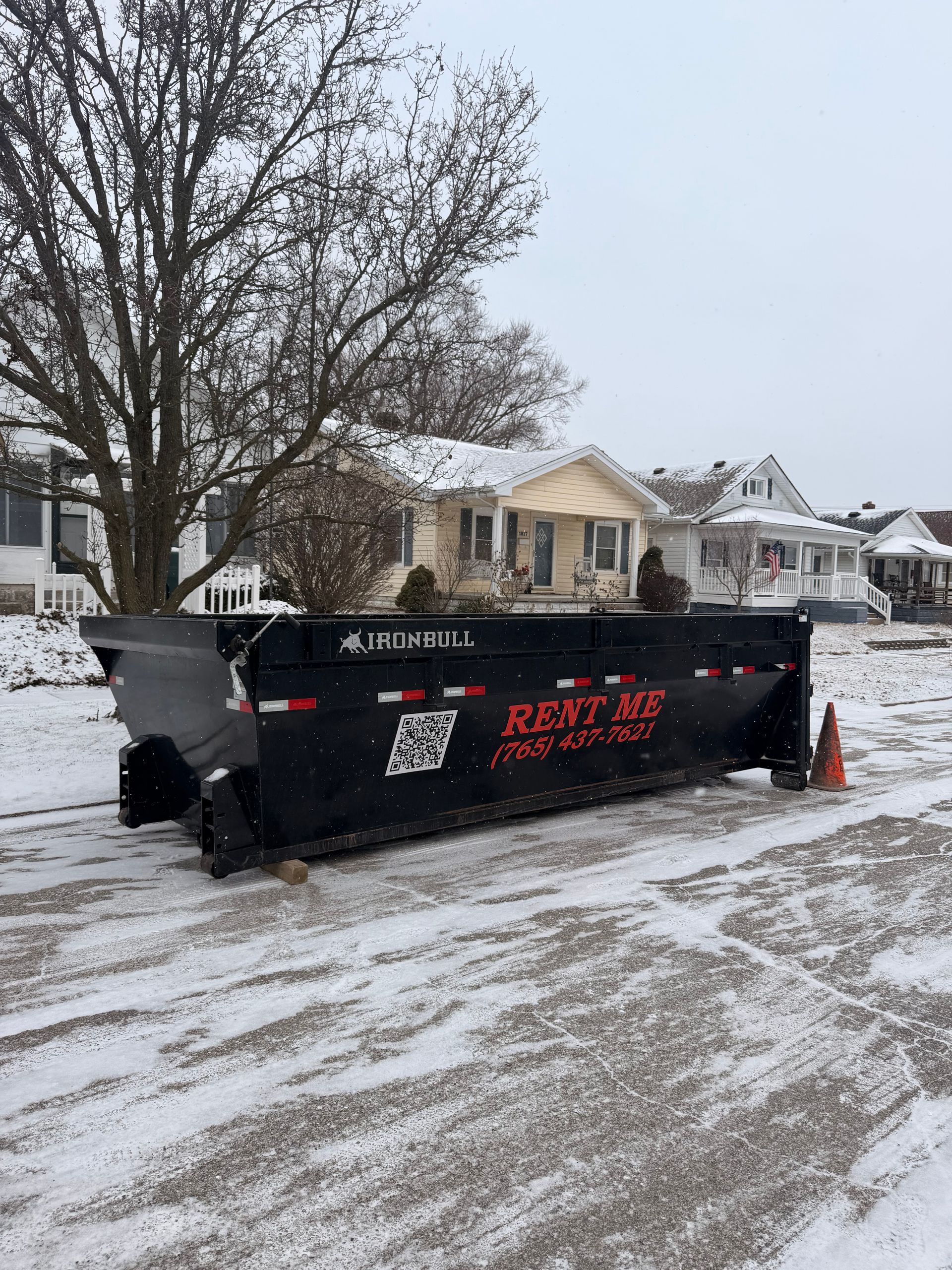 A black dumpster sits on a snowy residential street in front of houses, with 