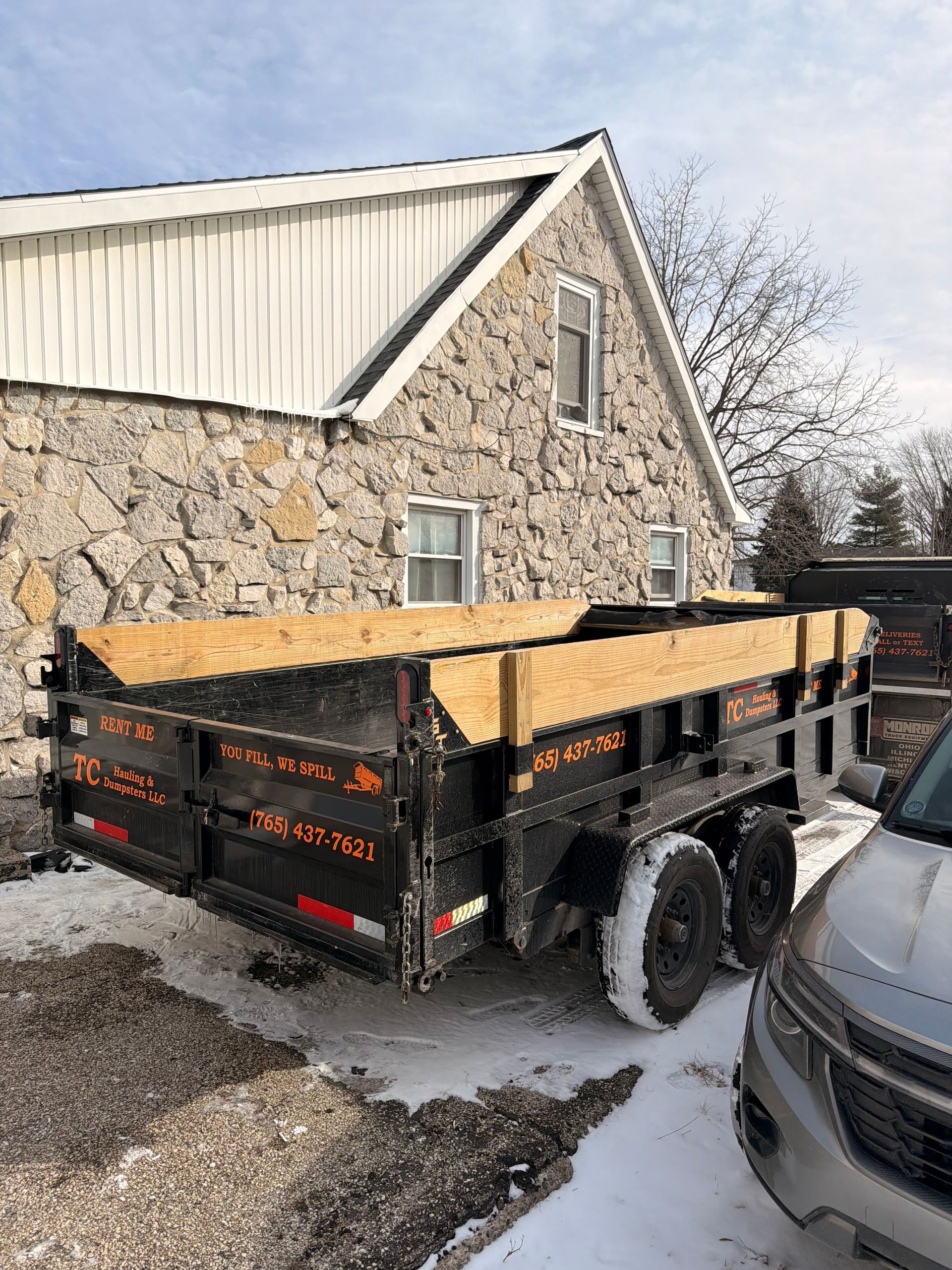 A black dump trailer with wooden side extensions parked on a snowy, gravel lot in front of a rustic stone building.