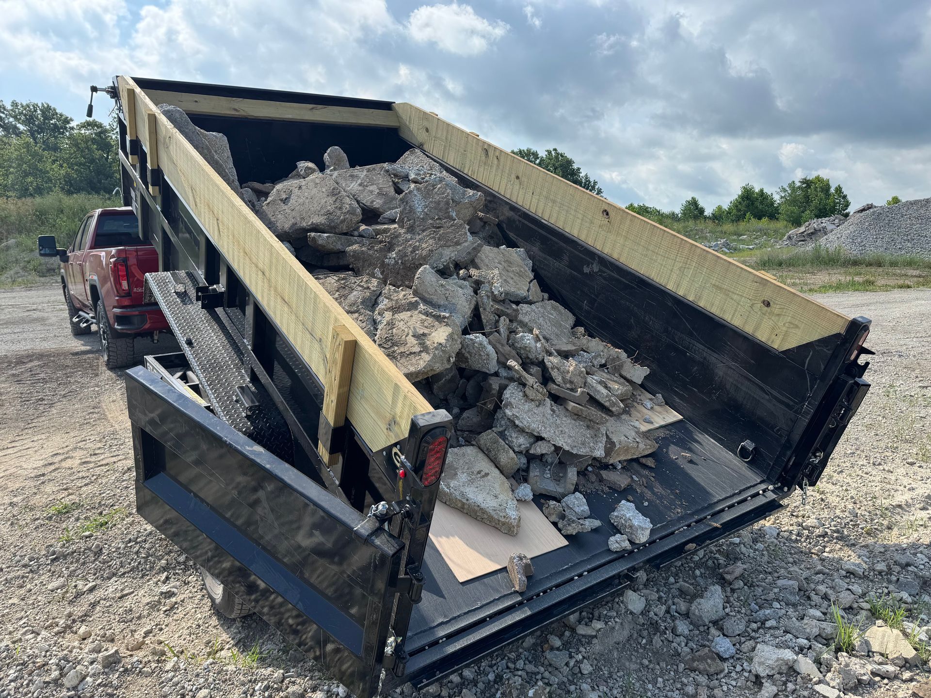A red pickup truck pulling a raised dump trailer filled with large chunks of concrete on a gravel lot.