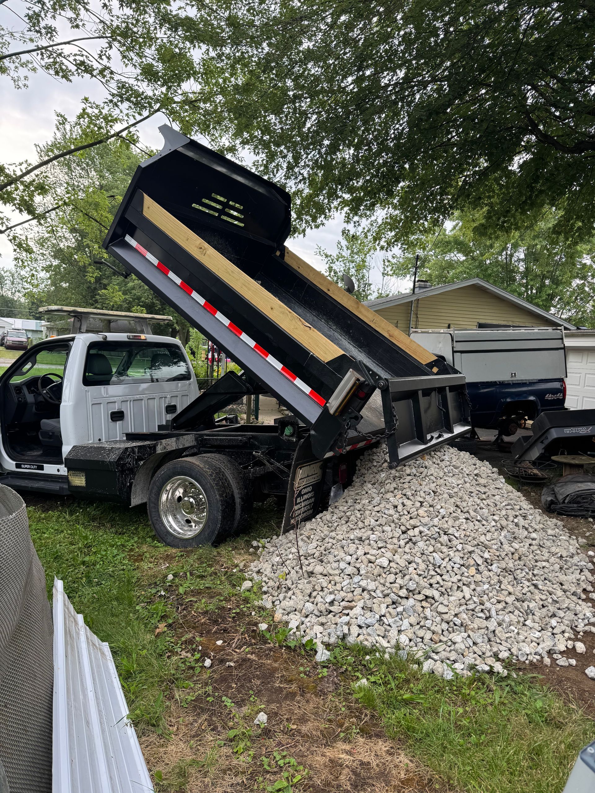 A white dump truck parked on grass with its bed raised, emptying a large pile of light-colored gravel onto the ground.