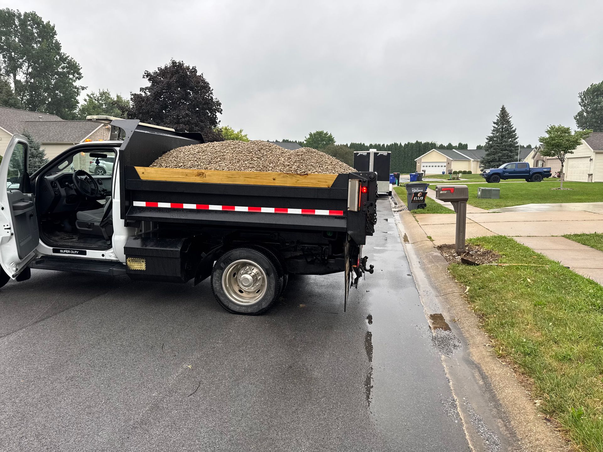 A white dump truck filled with gravel parked on a residential street during a cloudy day.