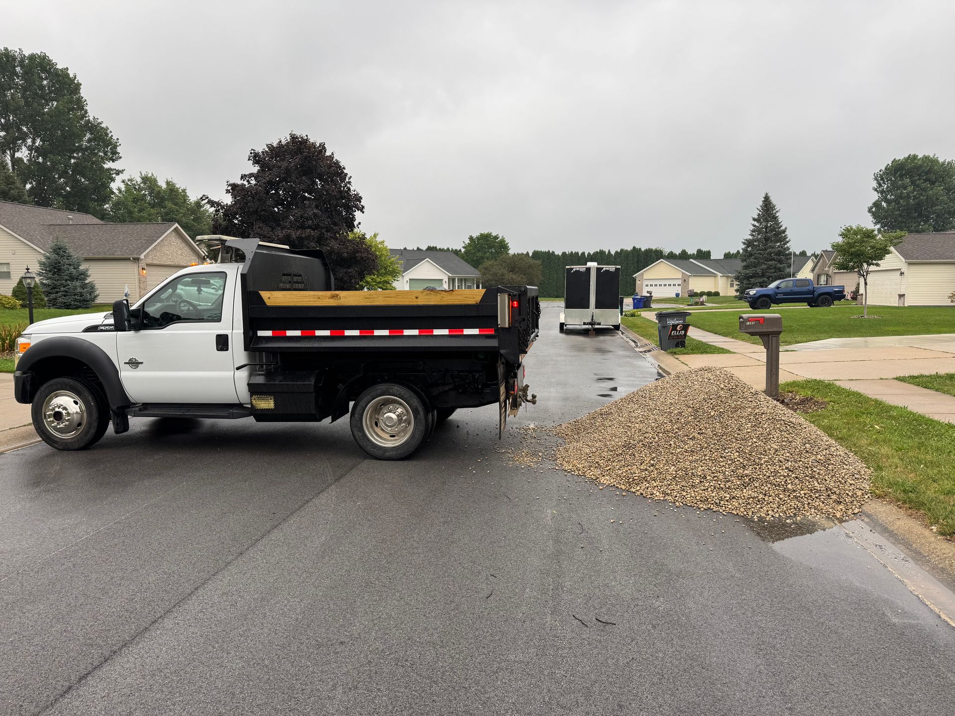 A white dump truck sits on a residential street next to a large pile of gravel deposited on the road.