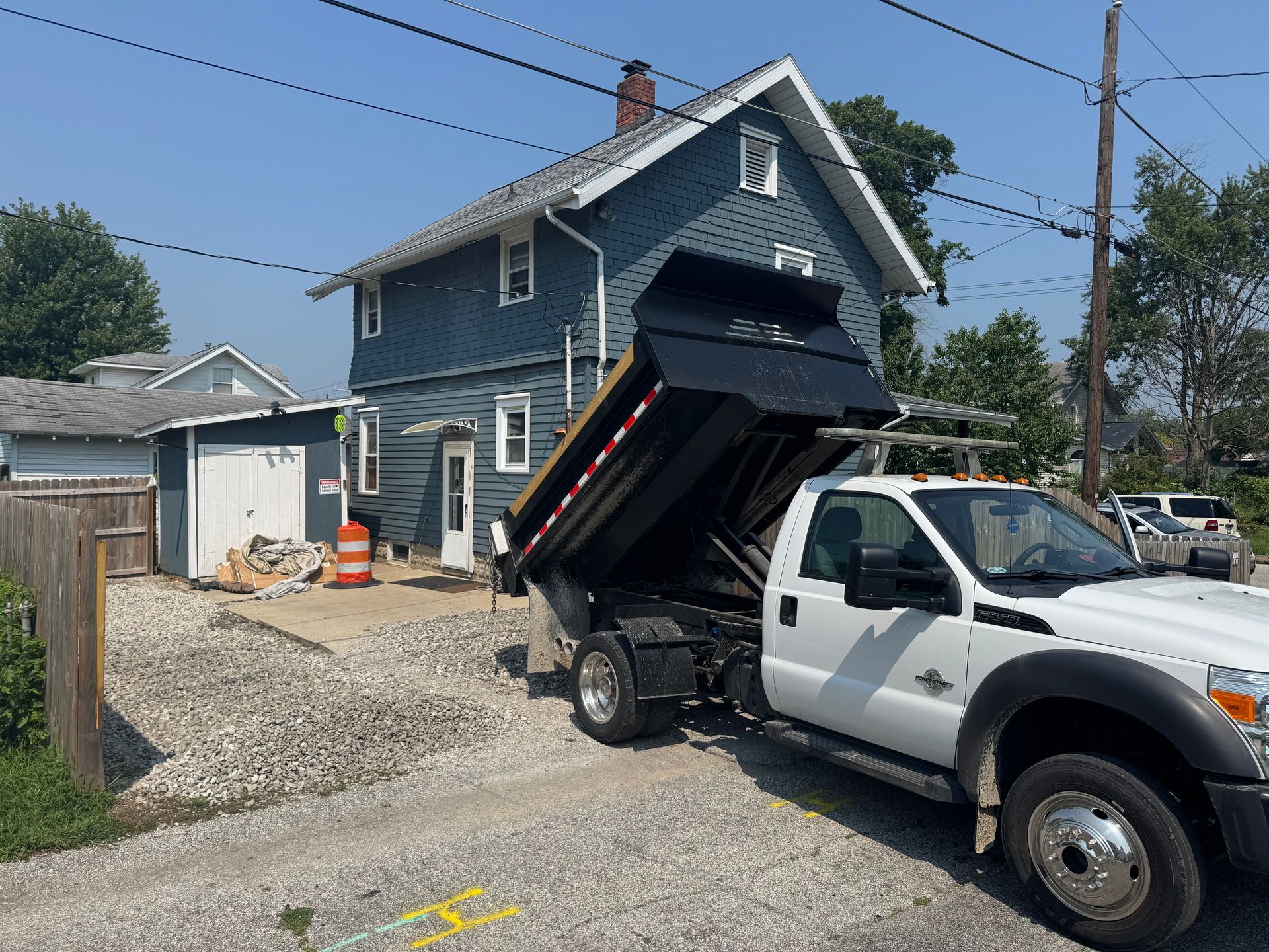 A white dump truck with its bed raised is parked on a gravel driveway in front of a blue two-story house.