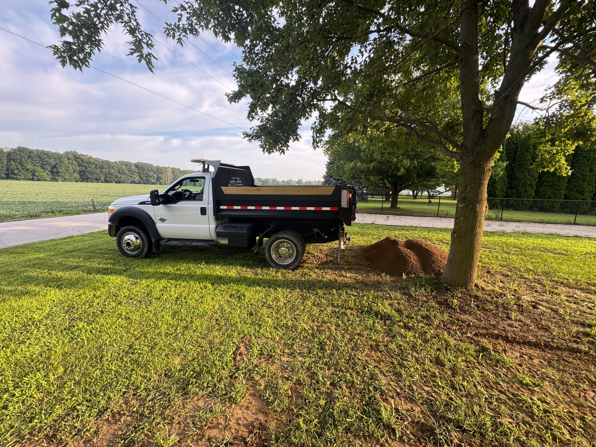 A white dump truck parked on a grassy field next to a tree and a pile of dirt, with a field in the background.