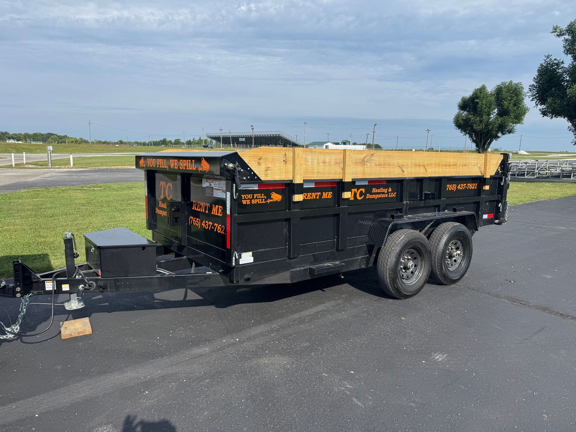 A black dump trailer with wooden side extensions parked on asphalt under a blue sky.