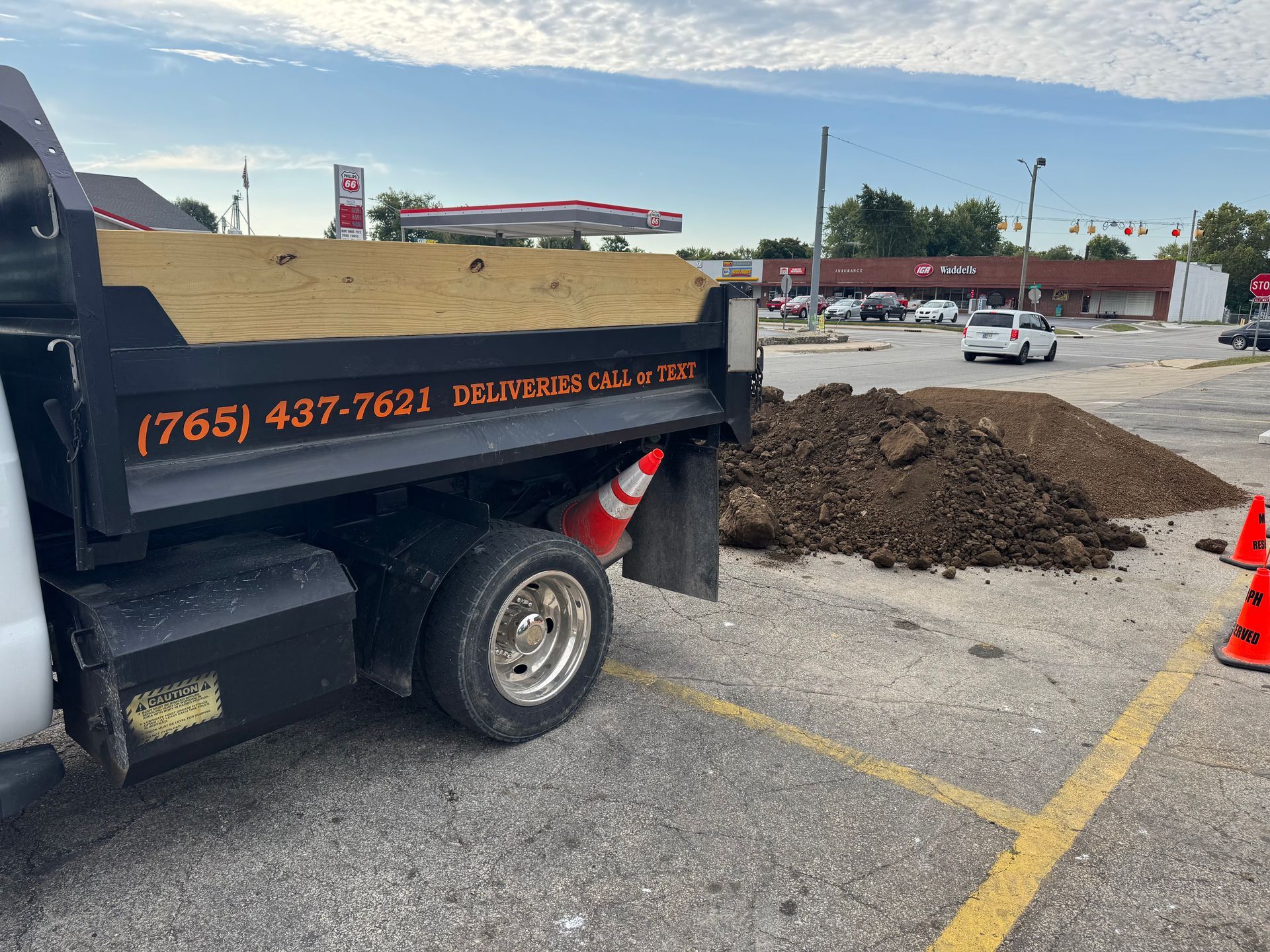 A black dump truck with wood-paneled sides parked next to a large pile of dirt in a parking lot with orange traffic cones.