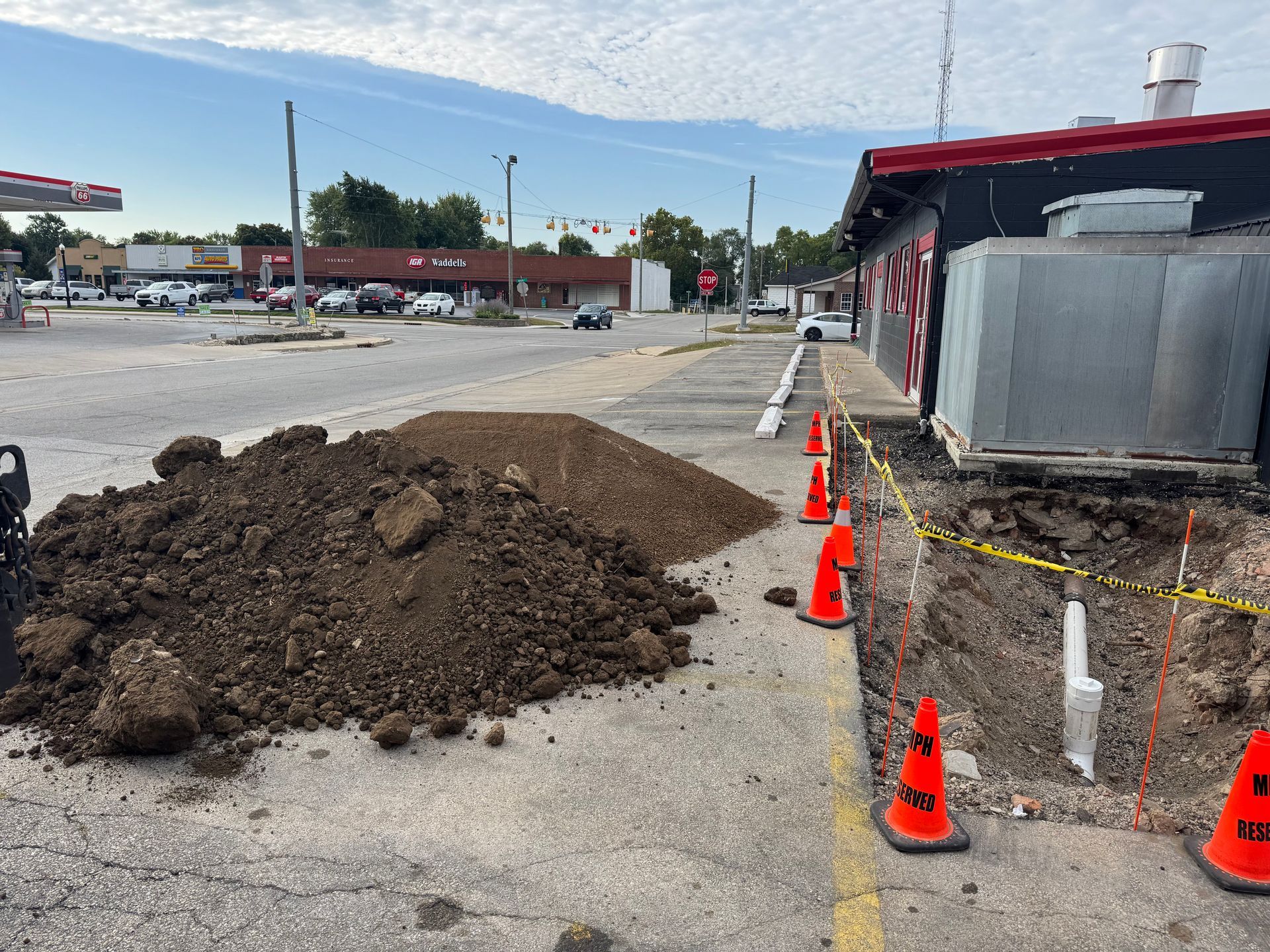 A large pile of dirt sits next to a construction trench marked by orange cones and yellow caution tape in a parking lot.
