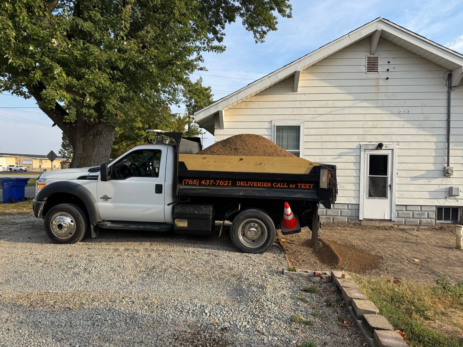 A white dump truck filled with gravel is parked next to a white house, unloading material onto the ground.