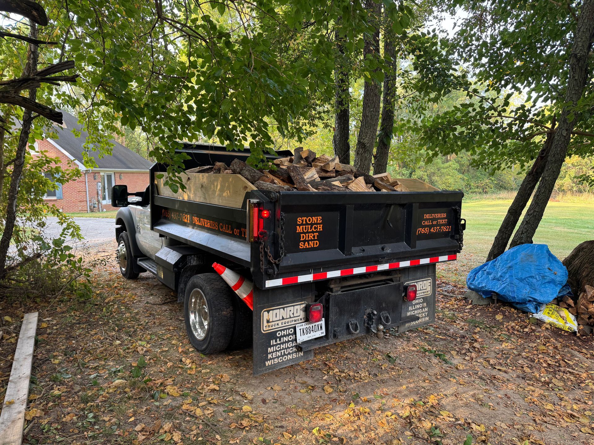 A black dump truck filled with firewood parked on a grassy, leaf-strewn area next to a house and trees.