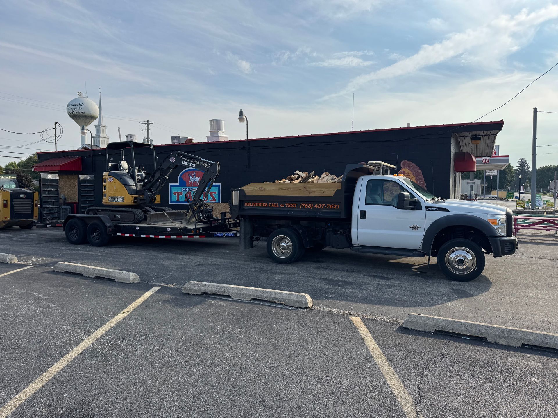 A white dump truck hitched to a trailer carrying a yellow CAT excavator parked in front of a black commercial building.