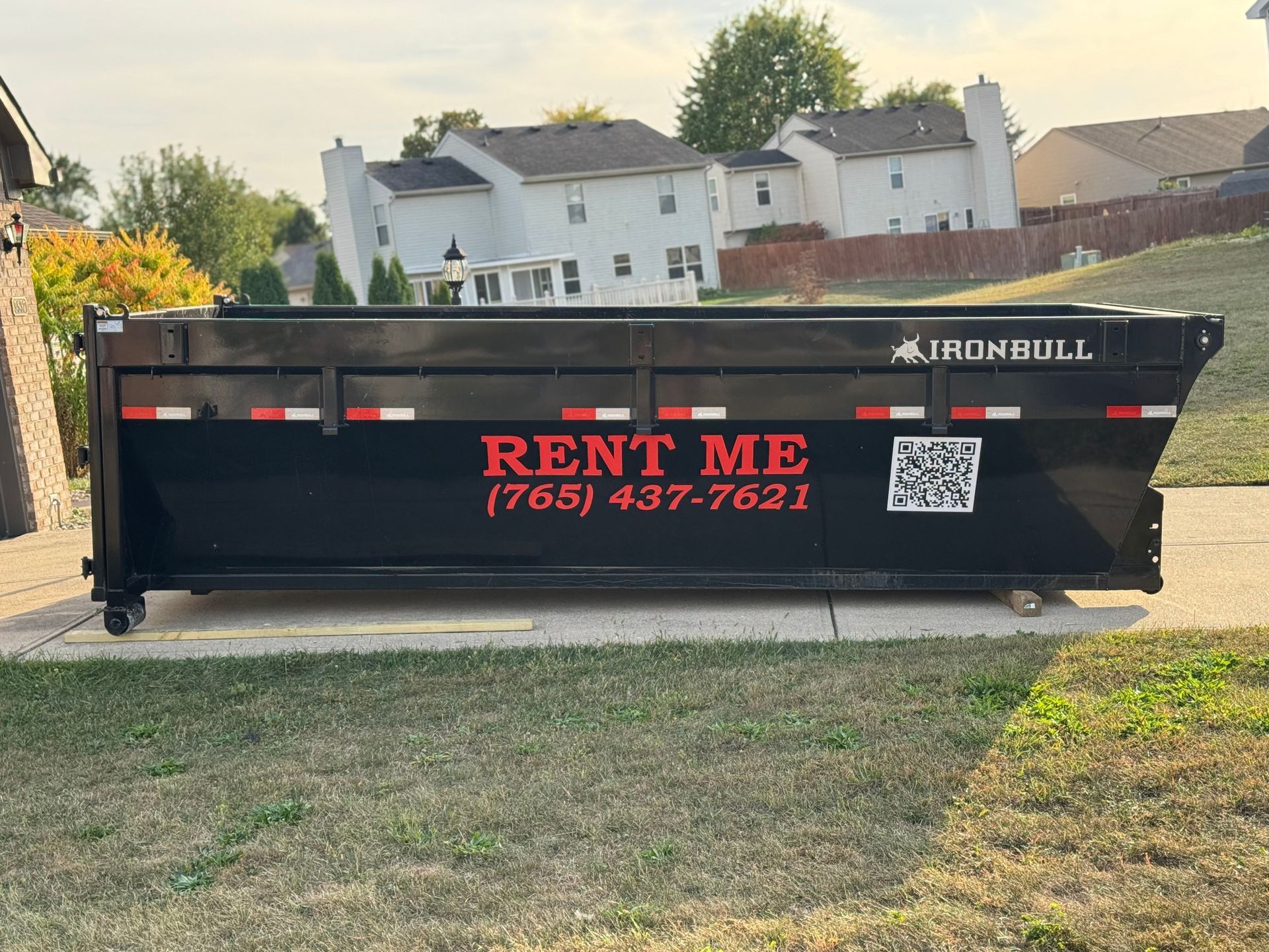 A black Ironbull dumpster for rent sits on a concrete driveway in front of a house, displaying a phone number and QR code.
