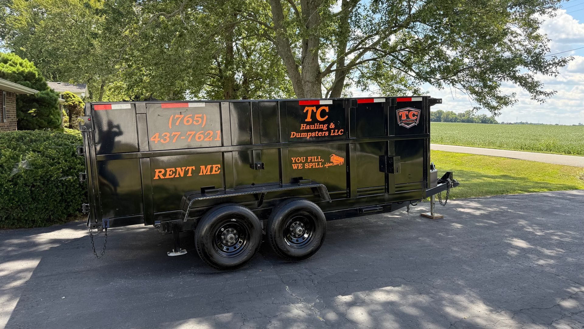 A black dumpster rental trailer parked on an asphalt driveway near green trees and a grassy field.