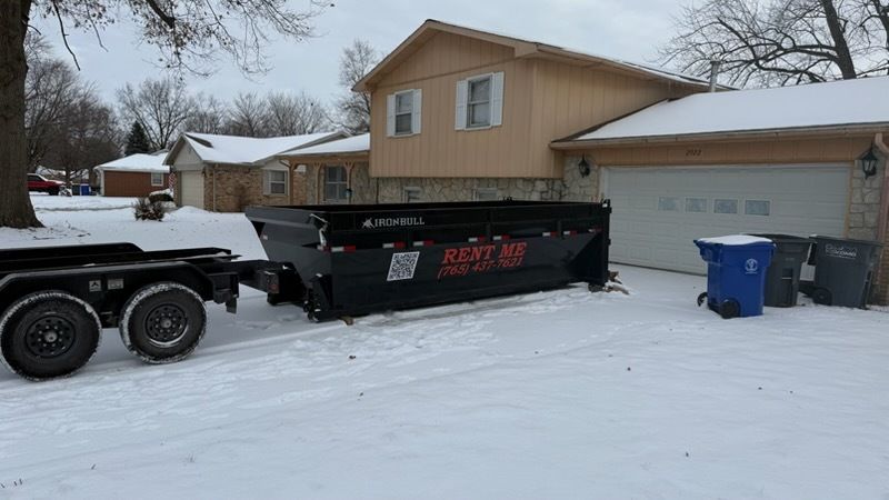 A red truck is towing a trailer full of logs in a grassy field.