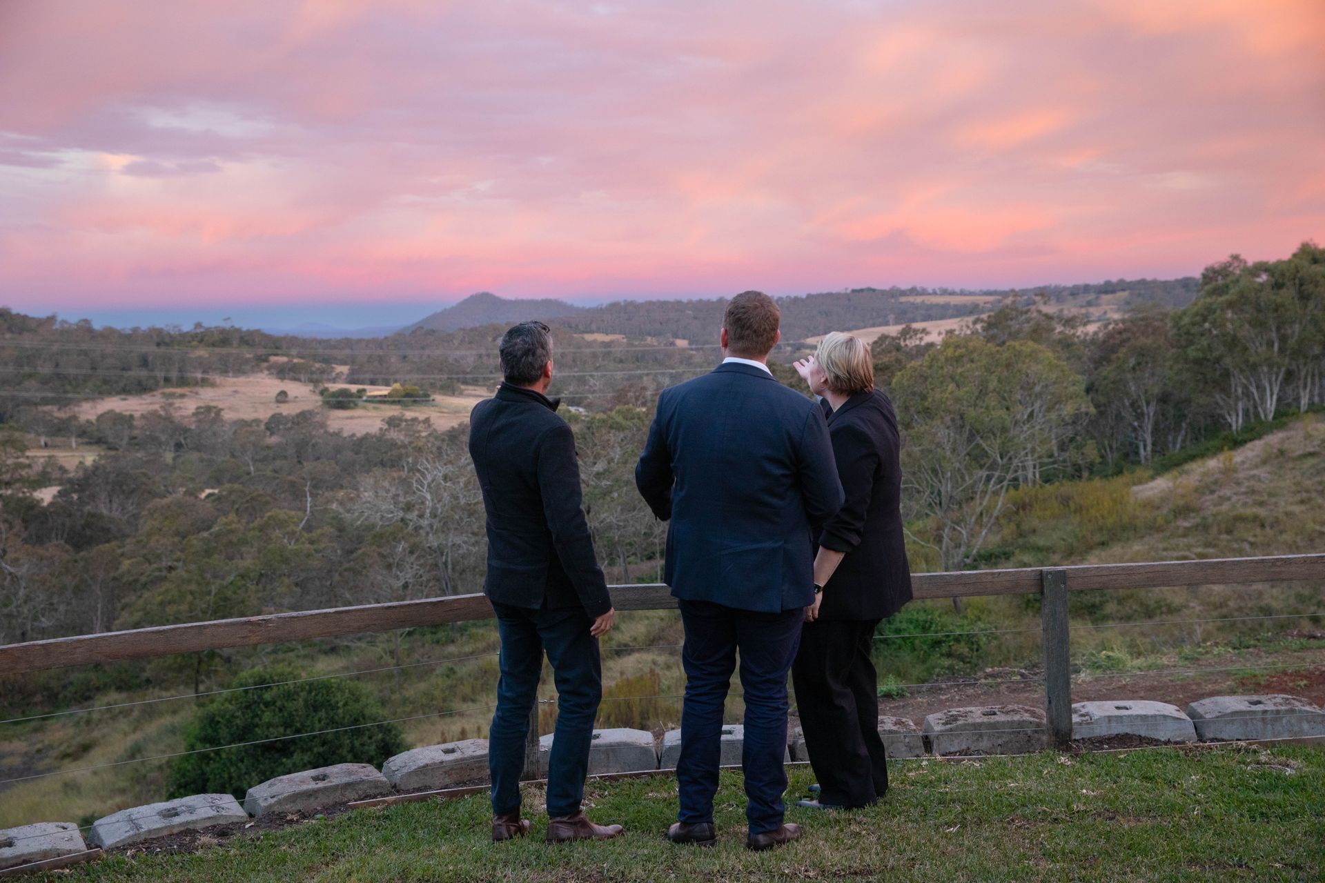 Three people stand on a grassy hill, gazing at a colourful sunset over a valley and distant ocean.