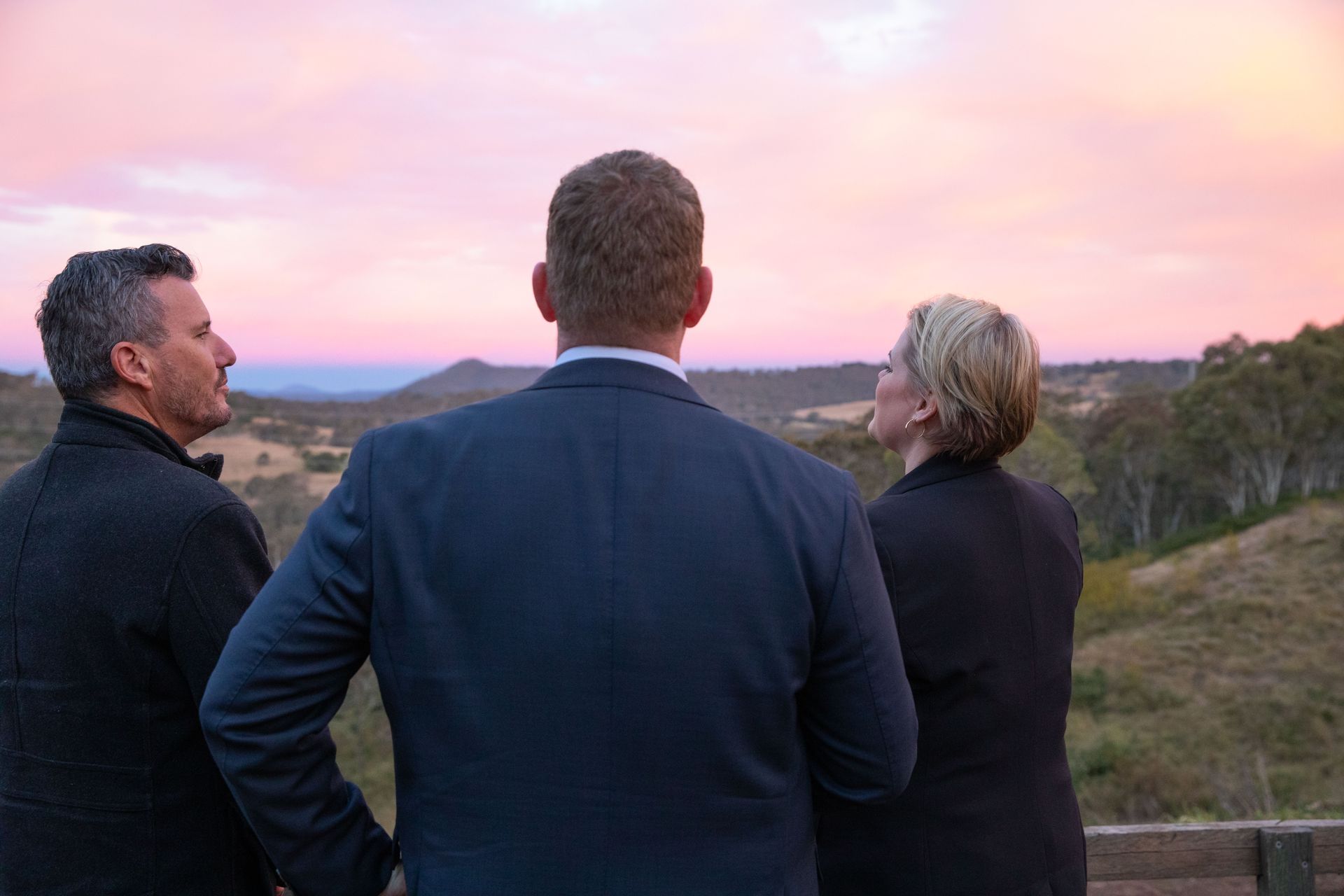 Three people in suits looking out at a scenic landscape at sunset. Pink and orange sky over rolling hills.
