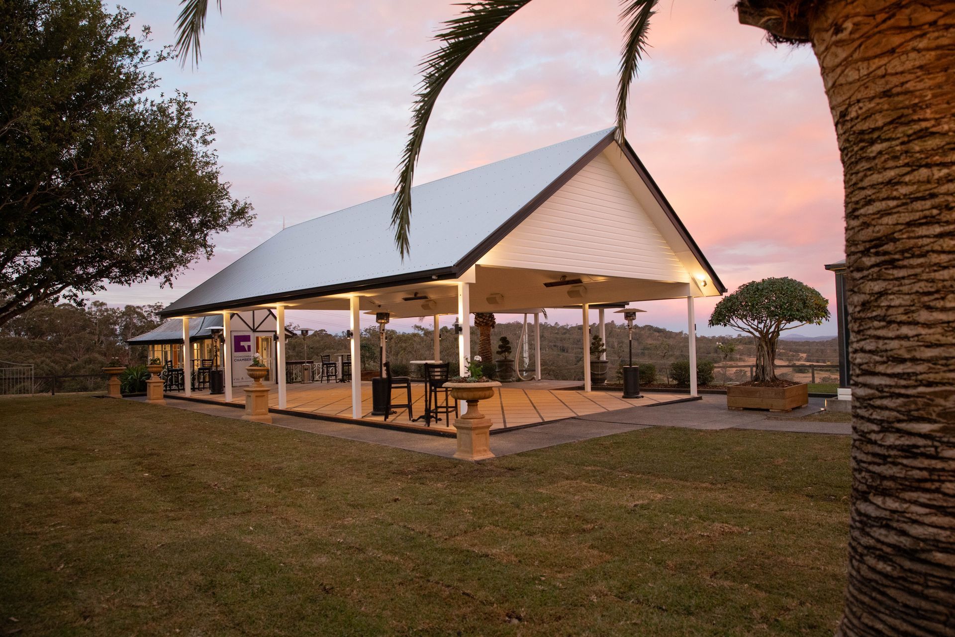 A pavilion with a white roof and wooden deck at dusk; overlooking a landscape of trees and hills.