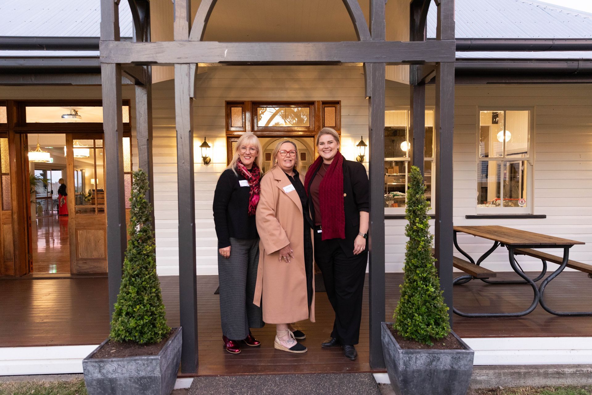 Three women smiling, standing on a porch framed by a dark wooden structure, in front of a white building at dusk.