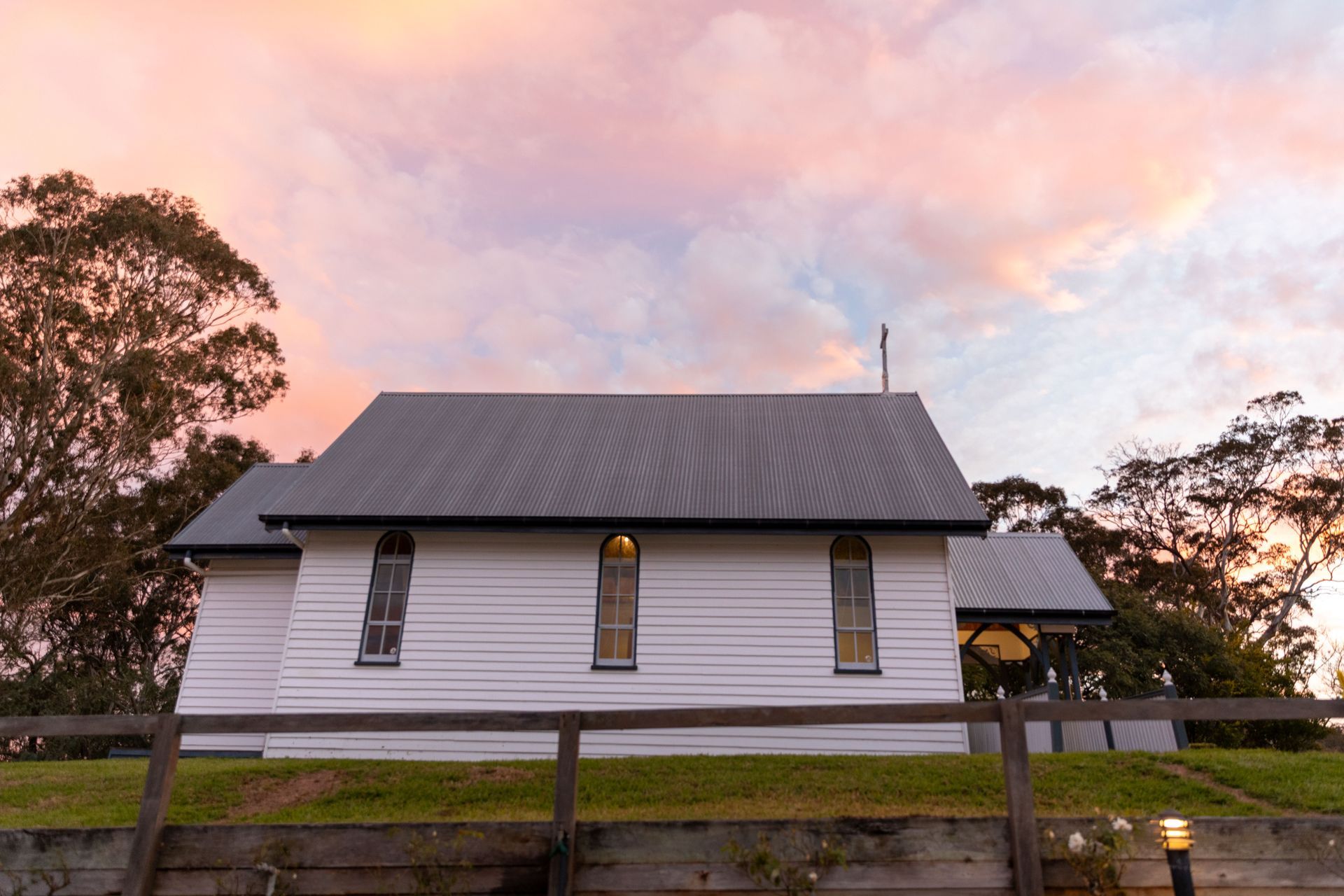 White church with a dark grey roof against a colourful sunset sky. The building sits behind a wooden fence.
