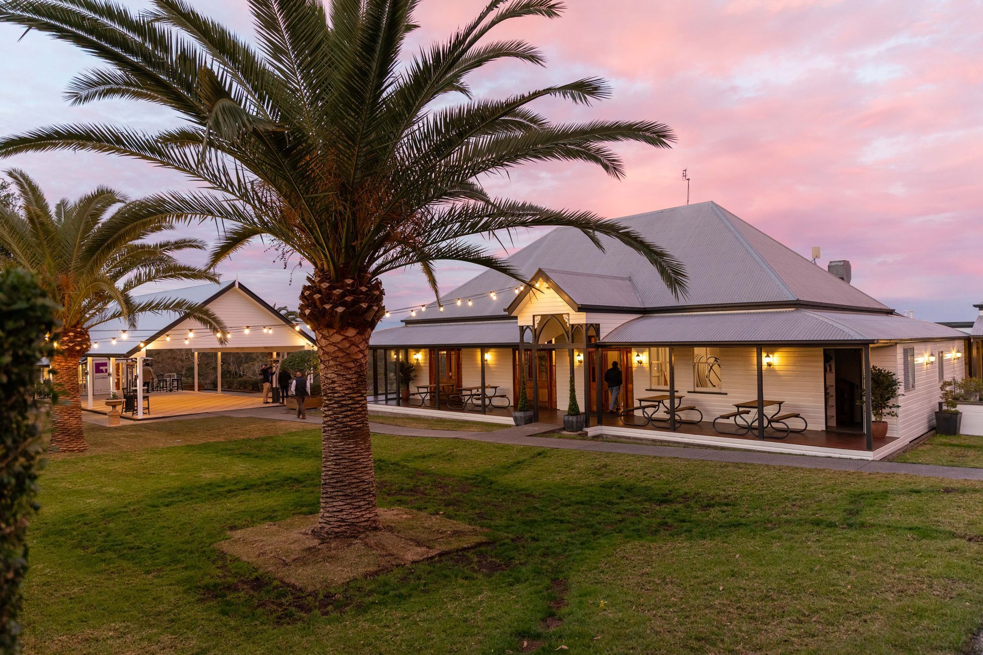 A long, white building with a wraparound porch under a sunset sky, palm trees in the foreground.
