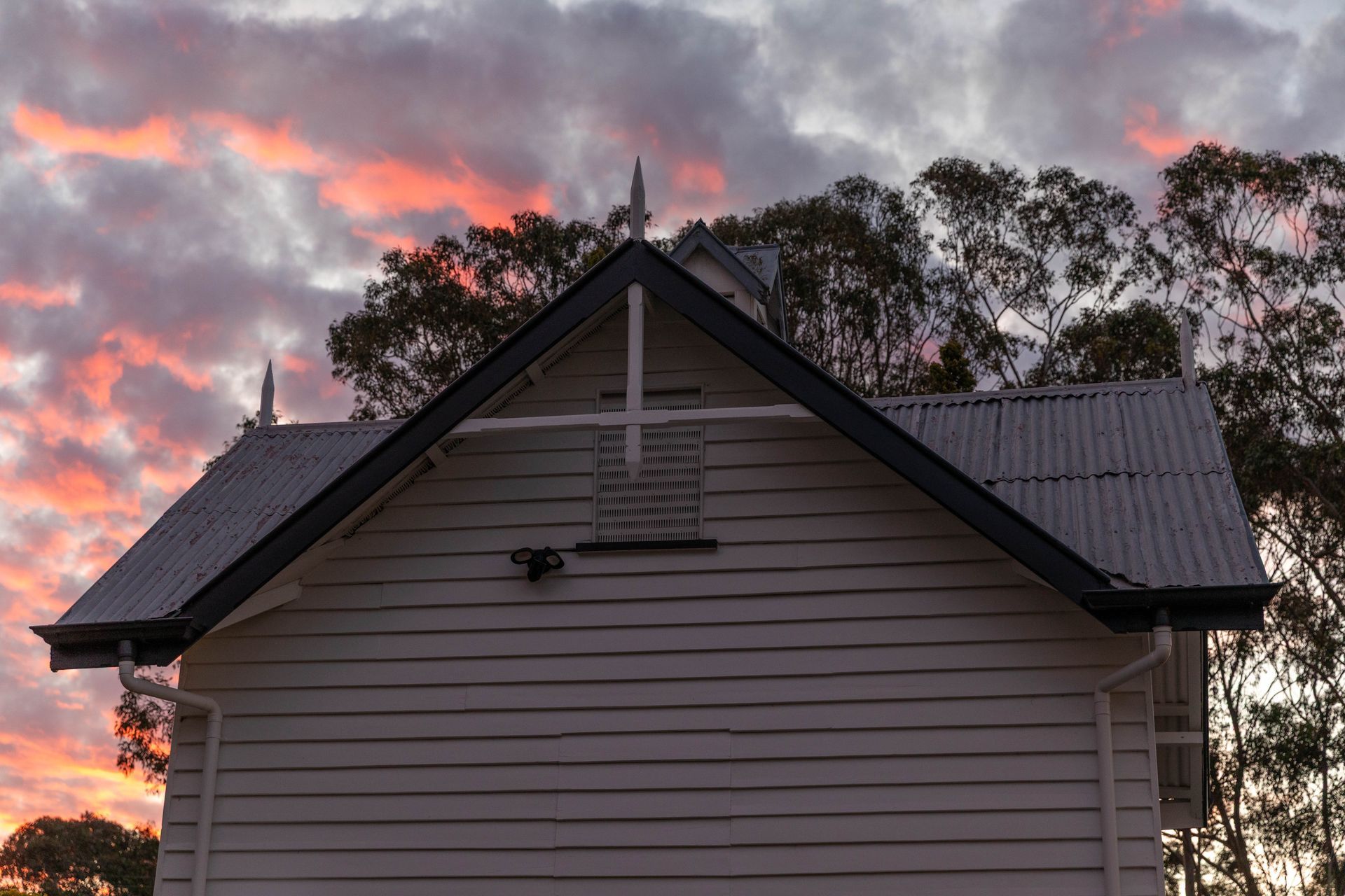 White building with a dark roof against a sunset sky of pink and orange. Trees frame the background.