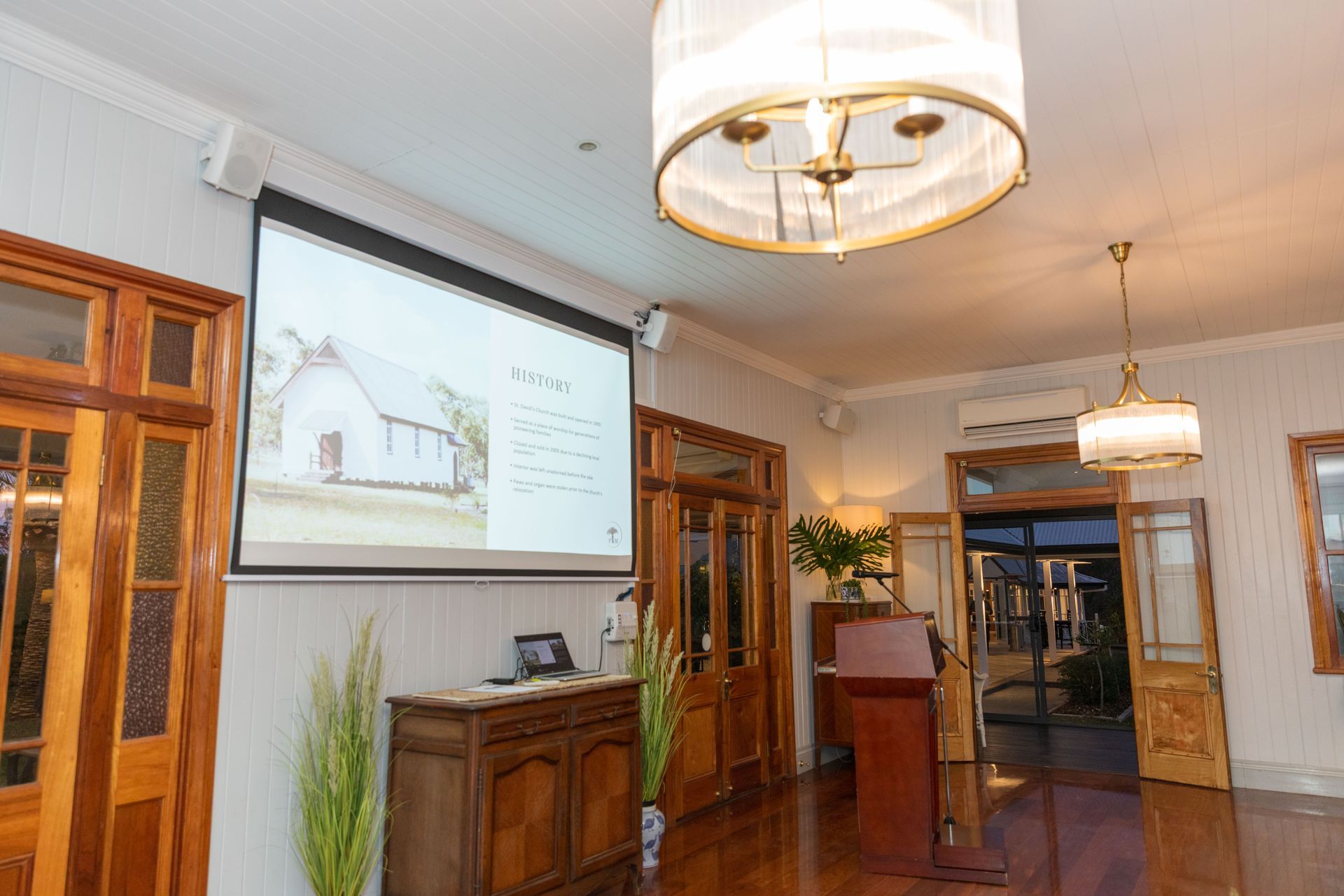 A presentation in a well-lit room, with a projector screen showing a building. Wooden doors and a cabinet are in view.