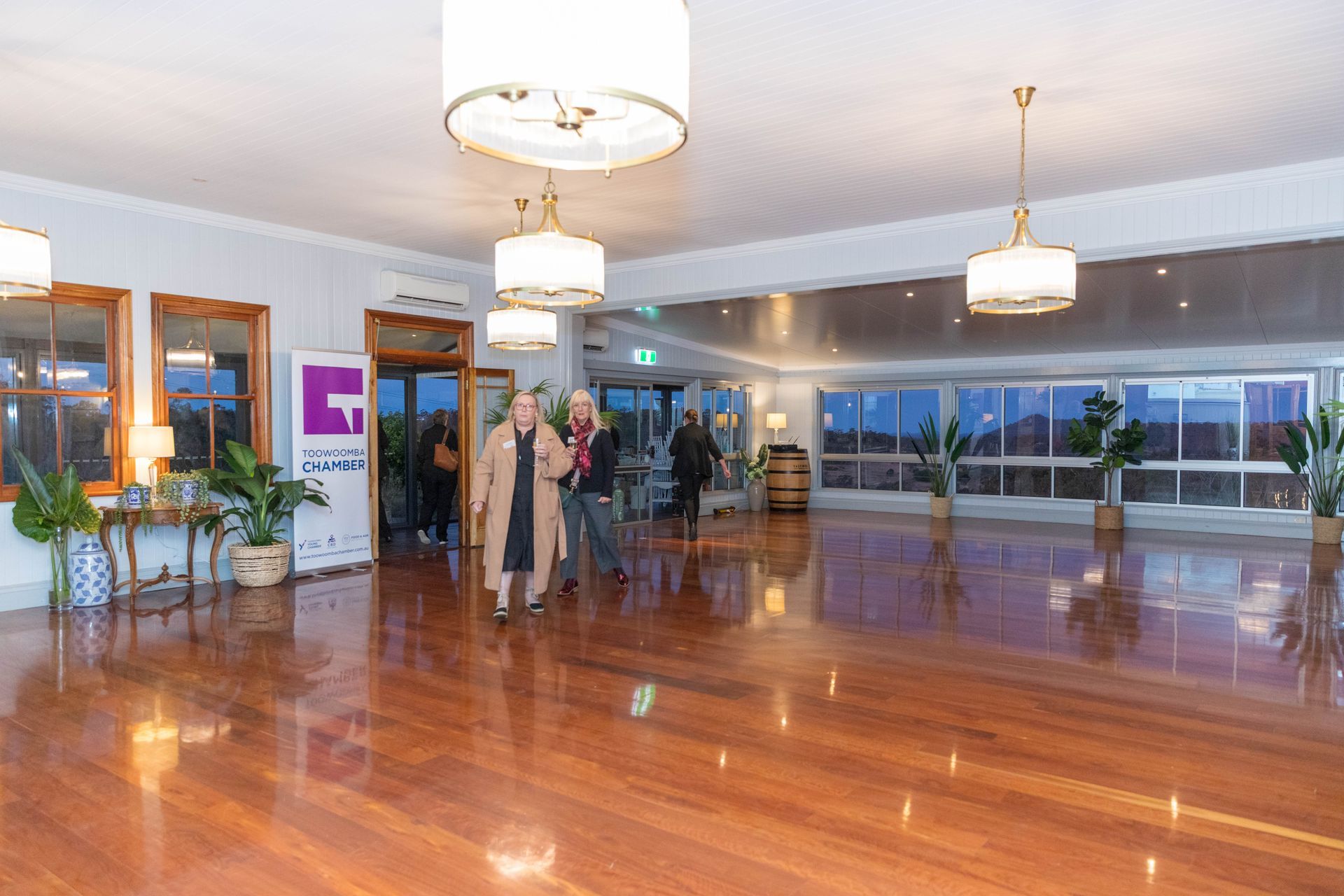 An empty event space with a wooden floor, large windows, and pendant lights. Two people walk toward the entrance.