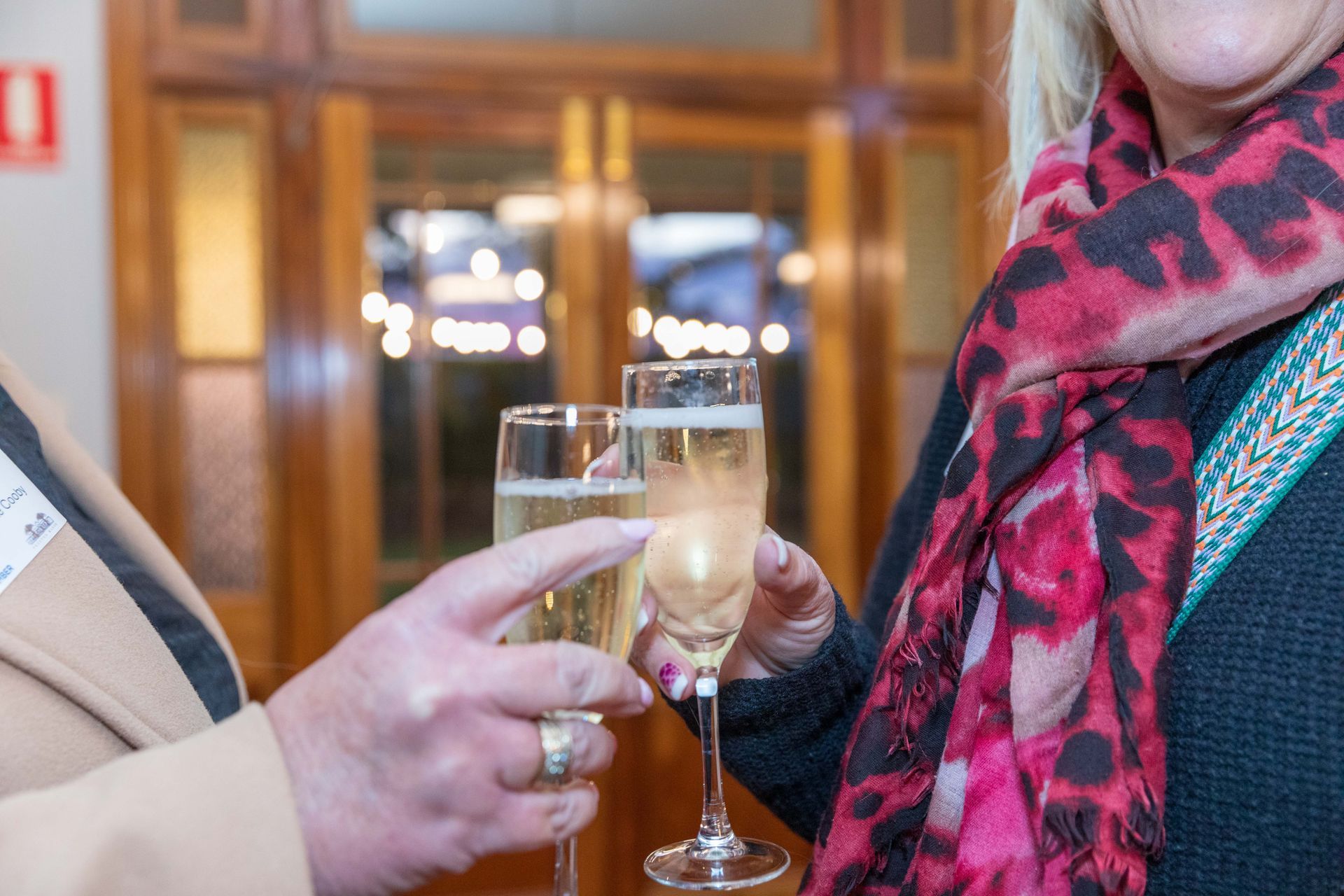 Two people clink champagne flutes, celebrating. One woman wears a pink and red scarf, the other has a name tag. The setting is indoors.