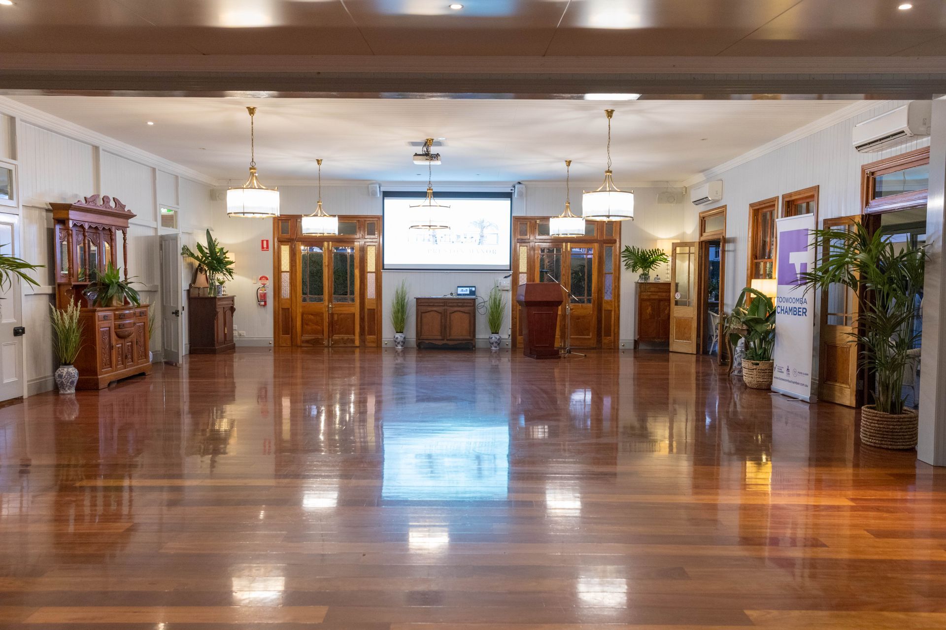 A spacious room with polished wooden floors and ornate wooden doors. A projector screen is centred on the back wall.