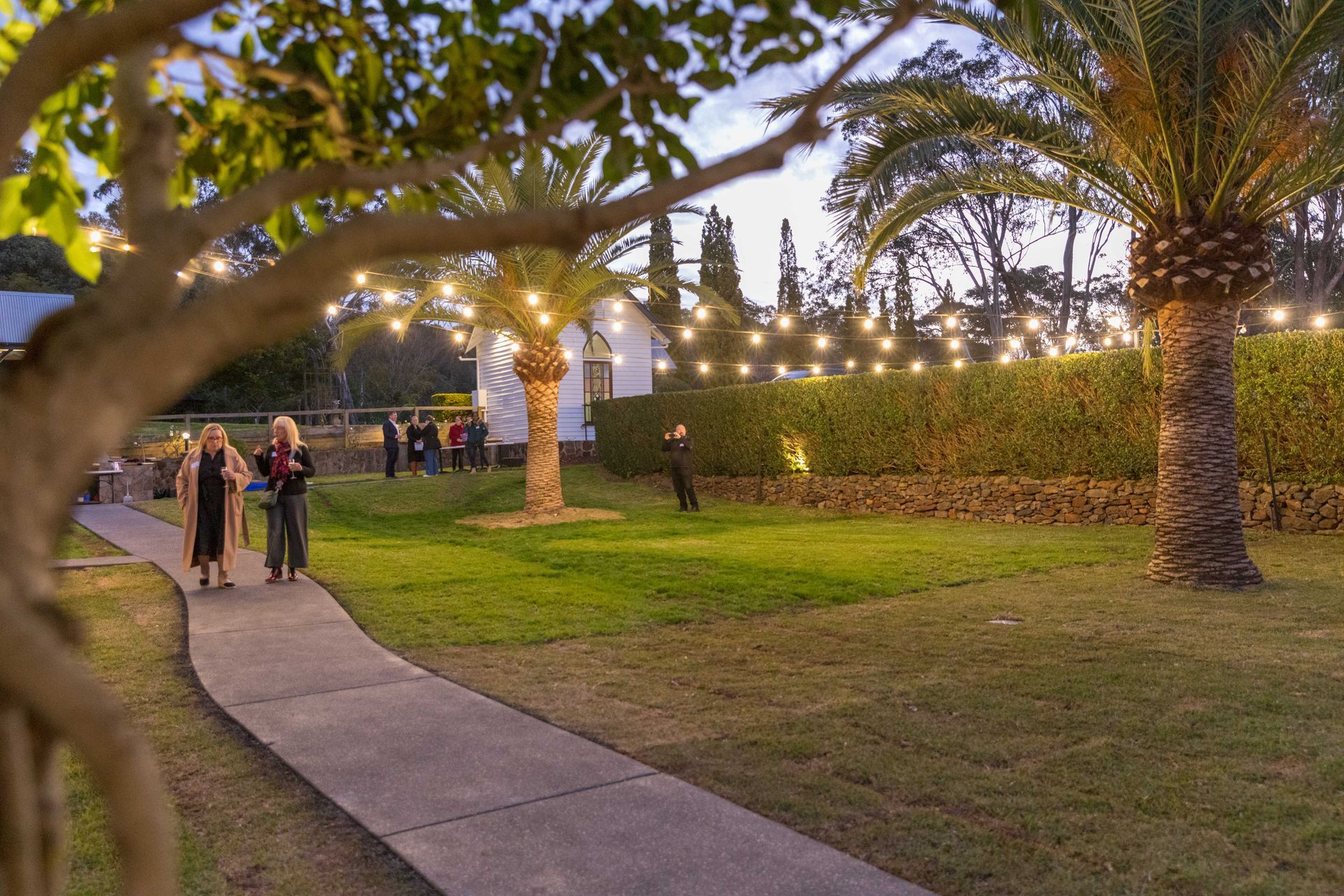 Two people walk along a path toward a building with string lights in a grassy yard at dusk. Palm trees and a hedge line the background.