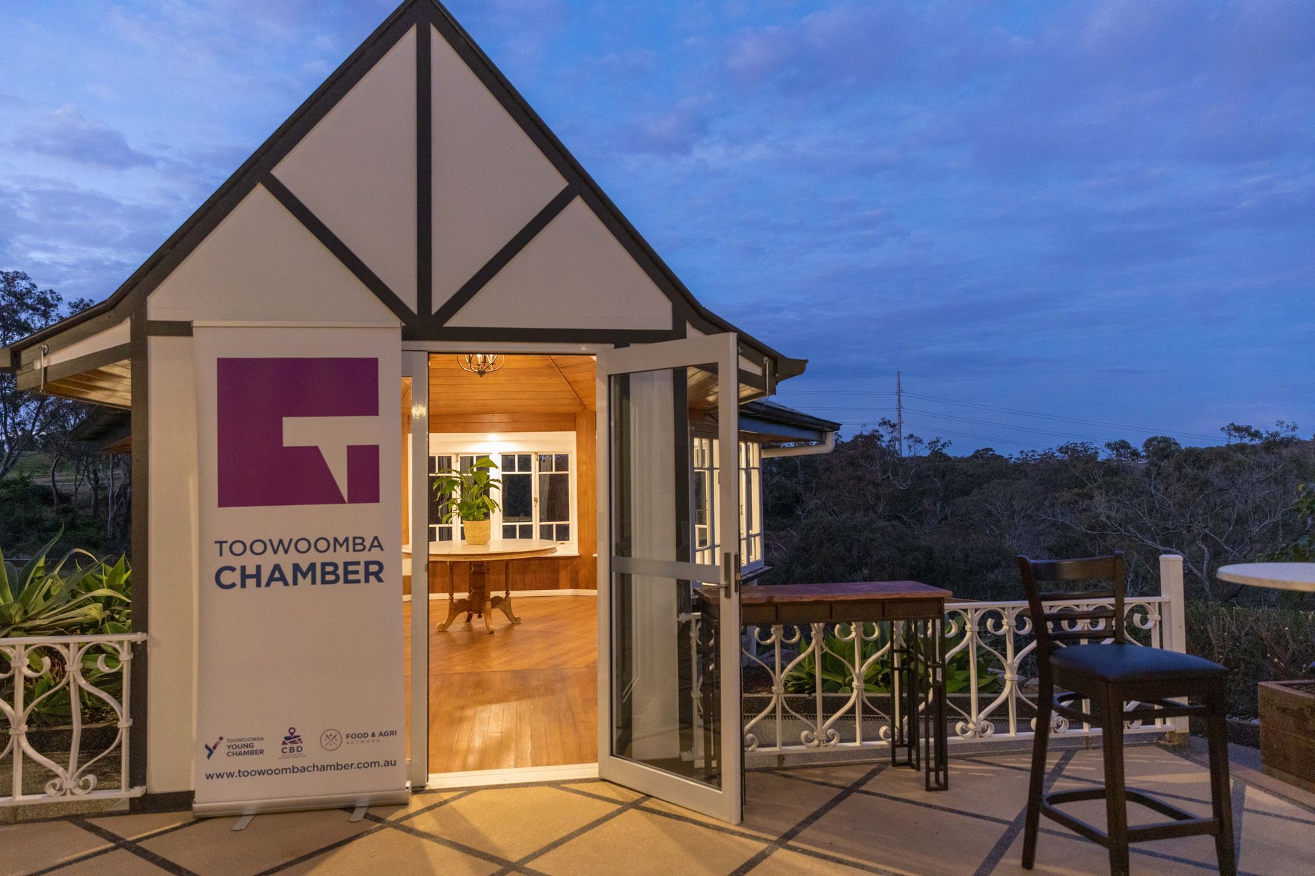 Small, gable-roofed building with an open doorway, displaying a banner for the Toowoomba Chamber. Overlooks an outdoor patio.